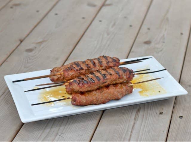A white tray holds kefta kebabs on skewers on a wooden table.