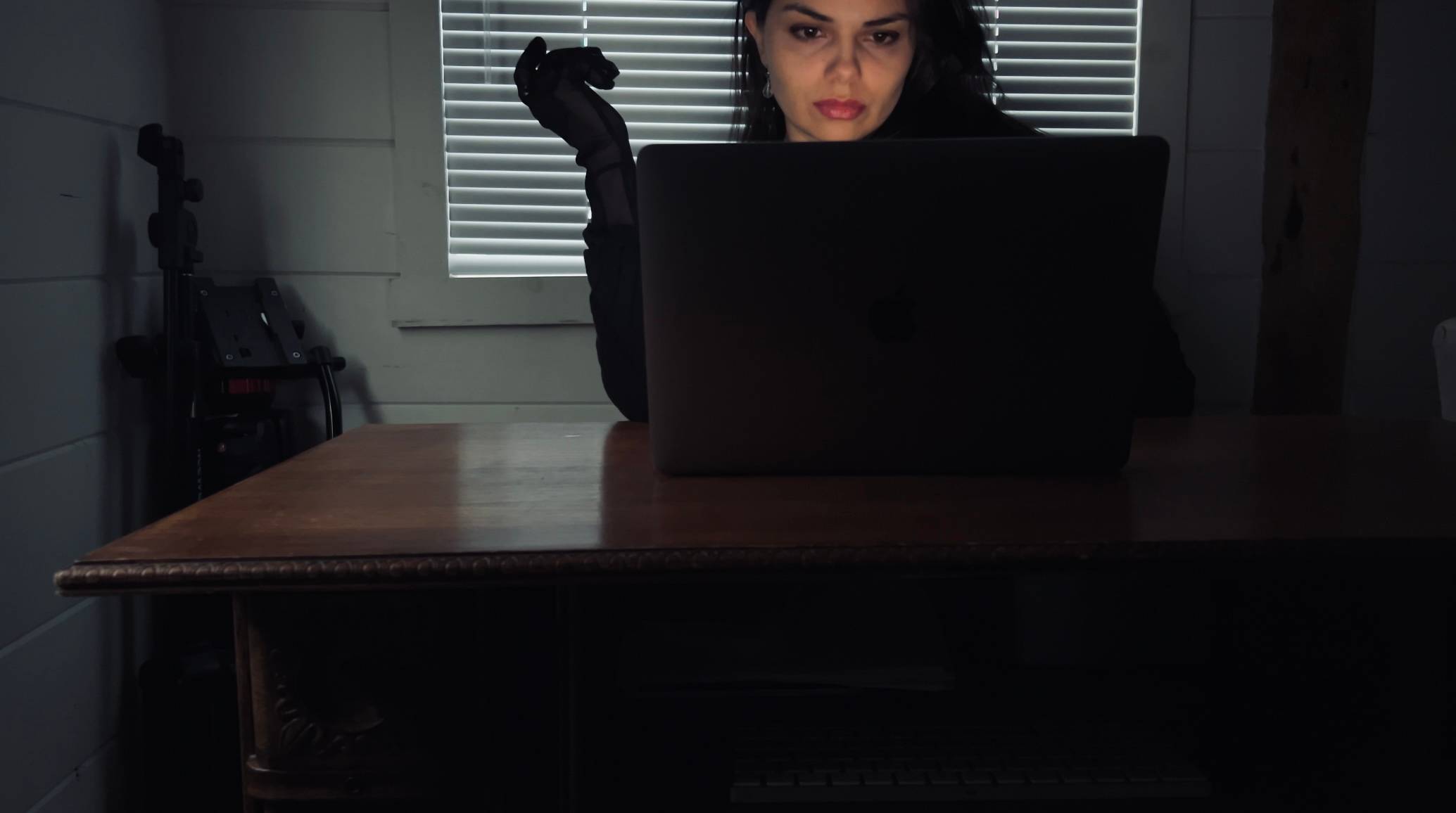 A noir-inspired image of Alina Medina sitting at her laptop in a dark office with the shades drawn, raising a gloved hand in thought.