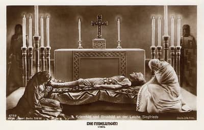 A black-and-white photo taken from a still from the Fritze Lang movie. Sifted is lying in state in front of a church altar, flanked by Brunhilde on the left and Kriemhilde on the right. 
