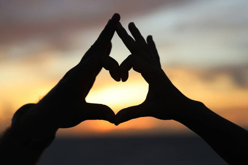 Silhouetted hands form a heart shape at sunset.