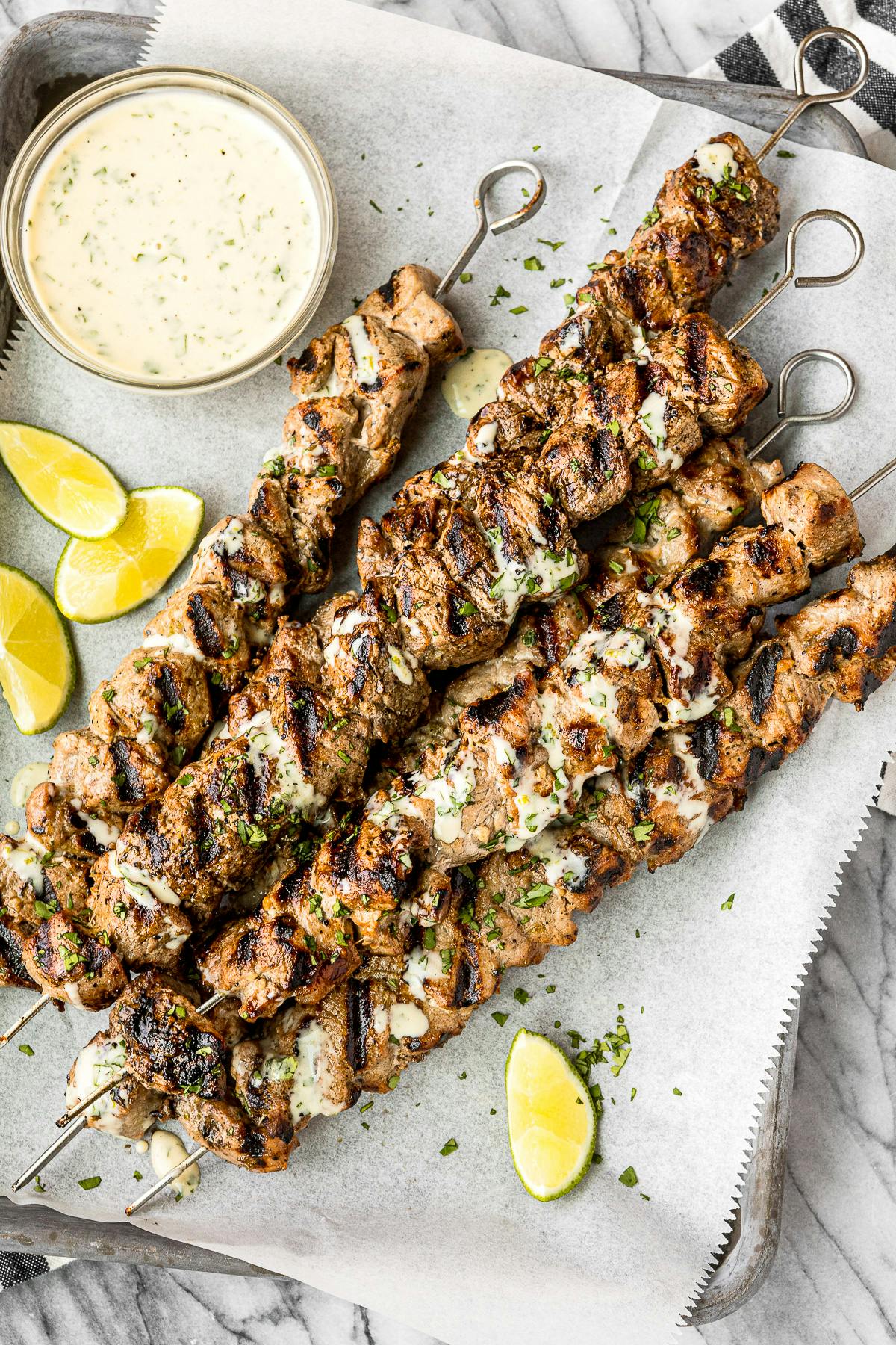 grilled pork kabobs on parchment paper with creamy cilantro slice and garnished with cut limes