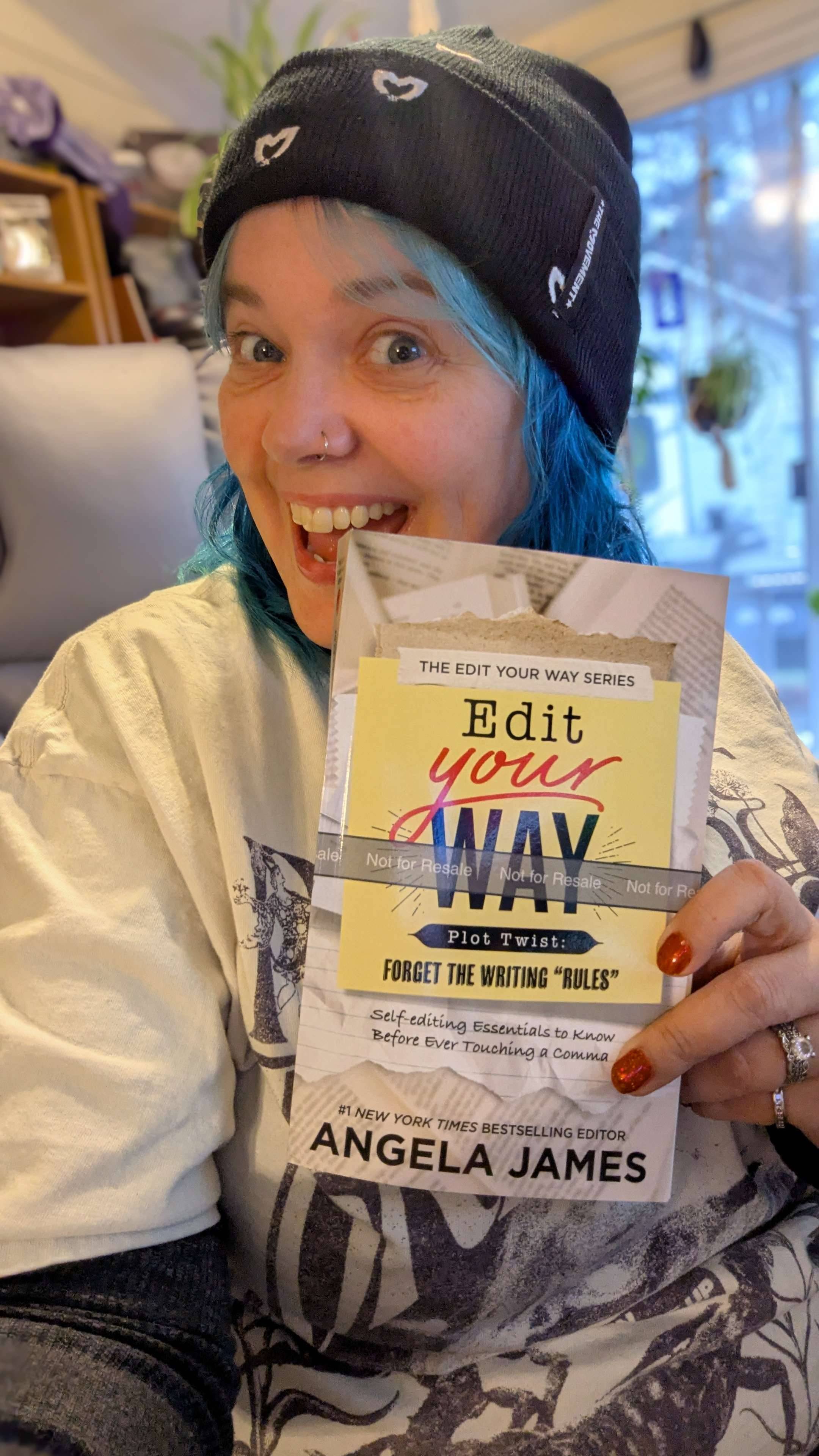 Angela James smiles excitedly while holding up the print proof of her book Edit Your Way. She’s wearing a black beanie with small heart-shaped eyes on it, and her blue hair peeks out beneath the hat. The cover of the book is clearly visible in the foreground, and the background shows a cozy indoor space with bookshelves and hanging plants.