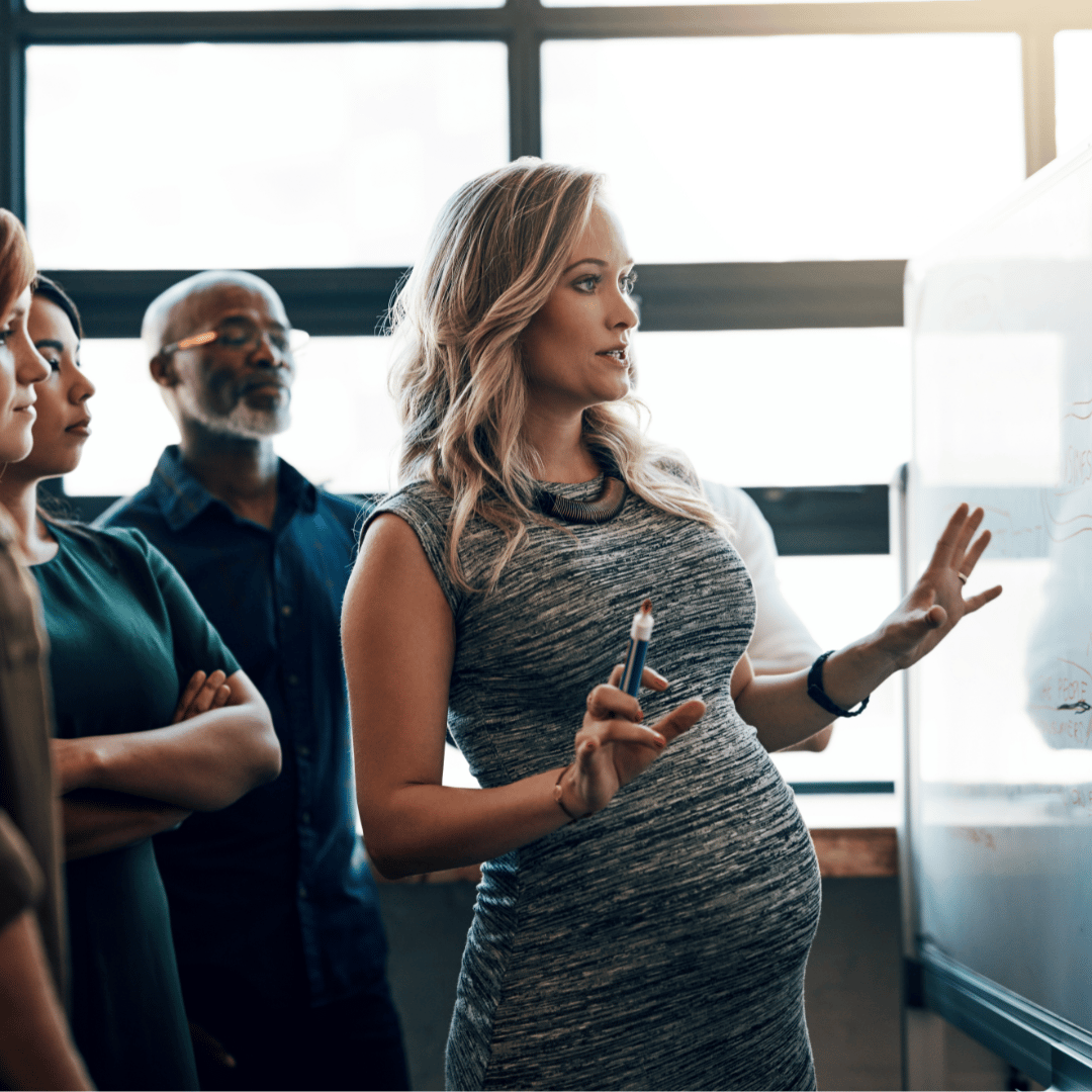 A pregnant woman leads a workplace discussion in front of a whiteboard.