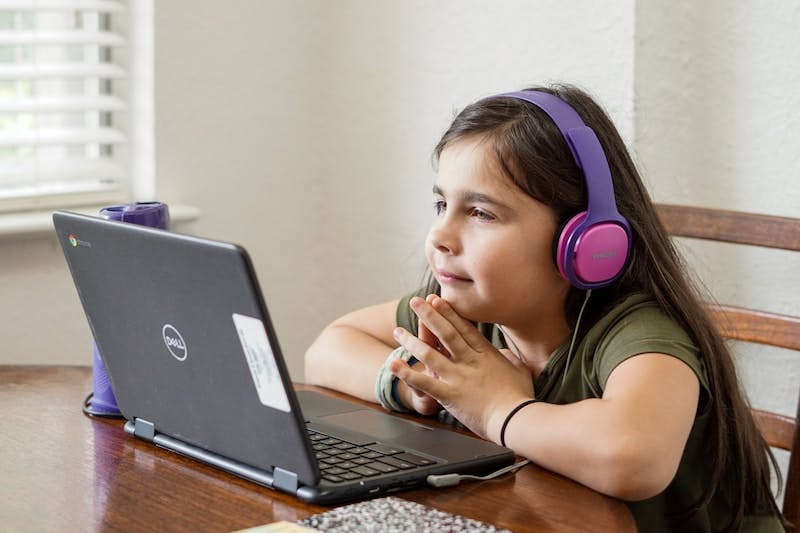 Young cute elementary aged girl with headphones on looking at a laptop while participating and learning in a remote virtual learning class during the COVID-19 pandemic quarantine.