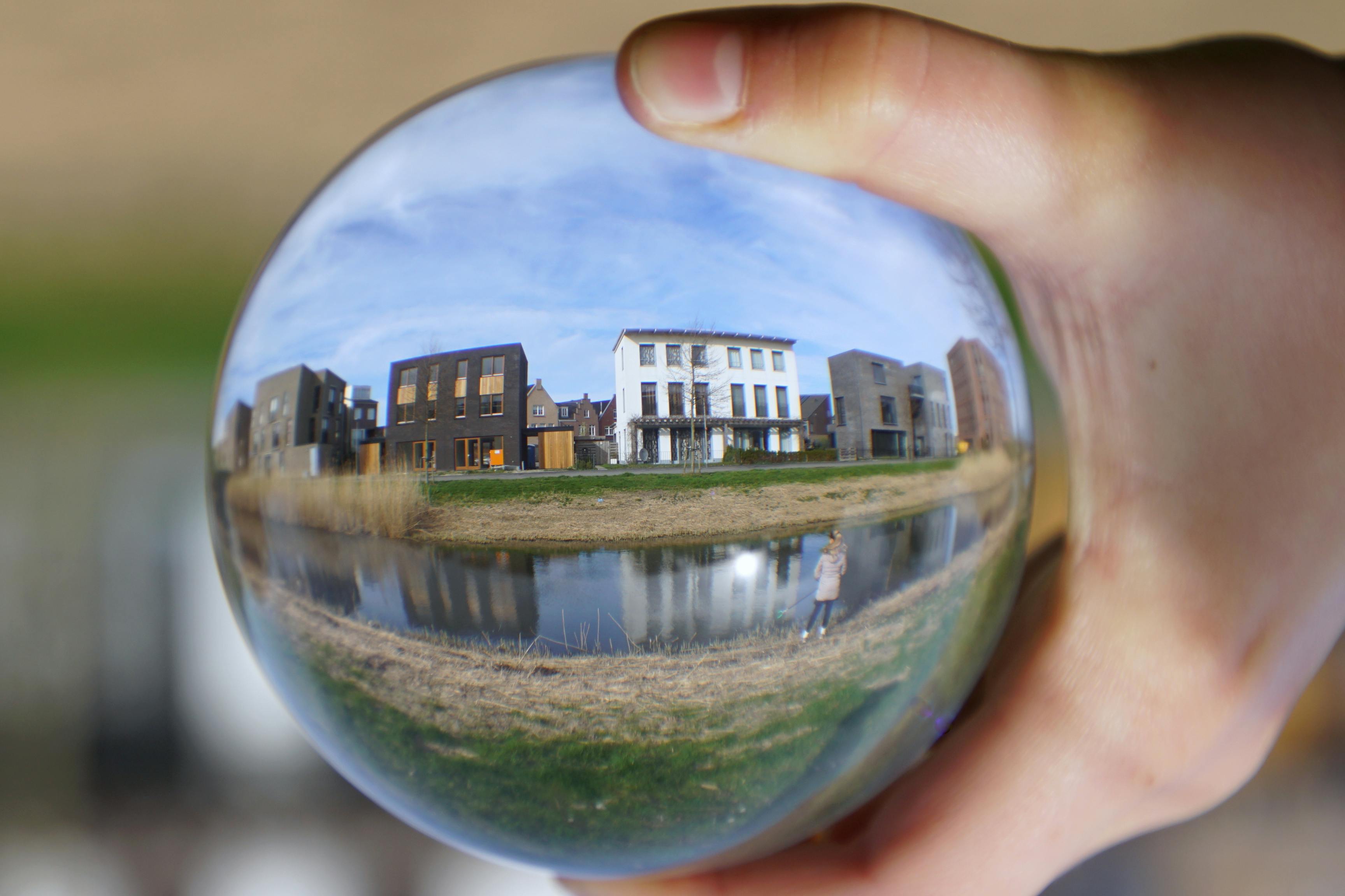 The image shows a hand holding a crystal ball.  Within the sphere are a row of three-story apartment buildings with one colored grey, one black, one white, and another grey. A blue sky with thin white clouds is overhead and in the foreground is a green and brown grass strip with a small canal running through the center of the area. The image of the crystal and housing conveys 7 predictions for the 2024 rental market.