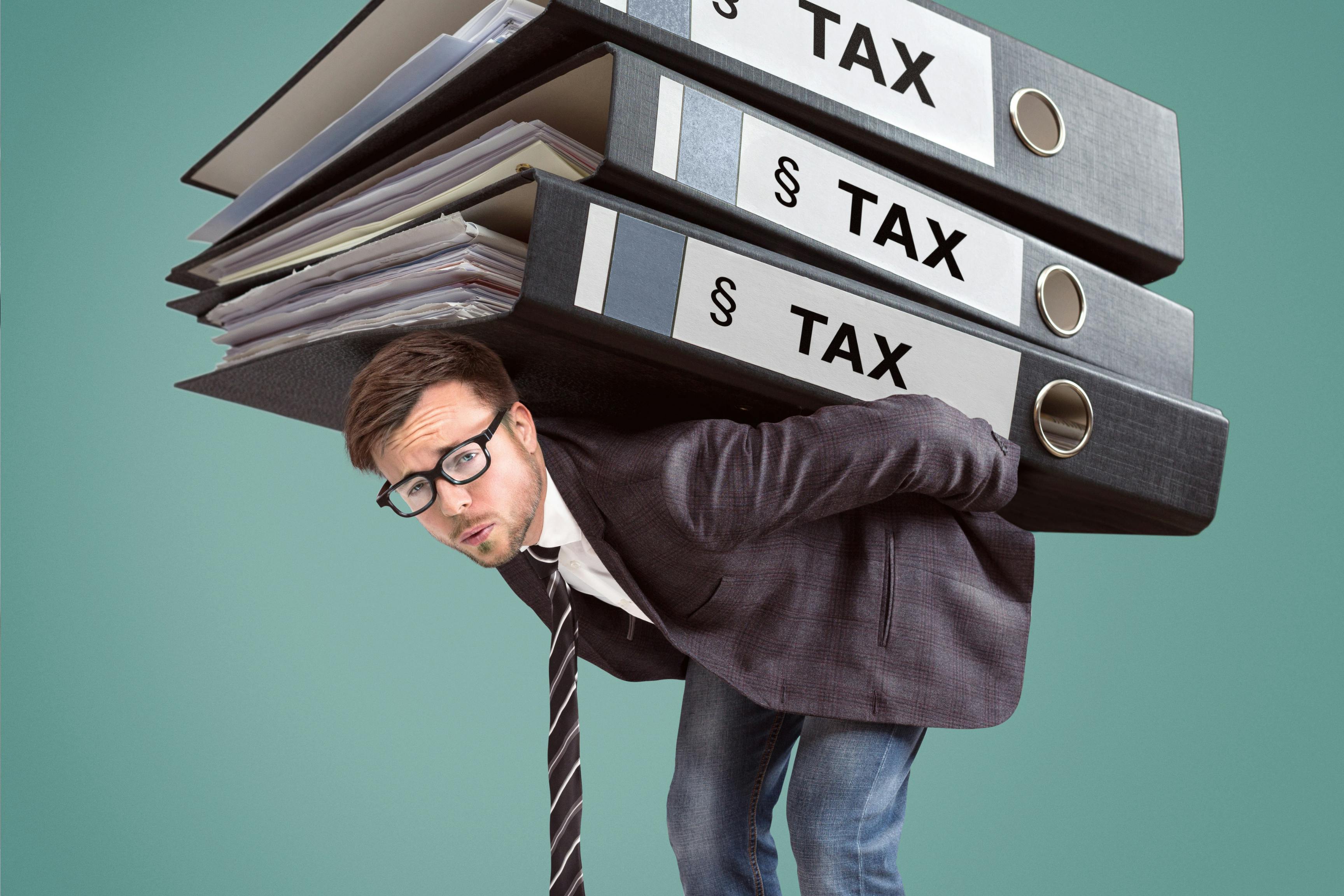 The image shows a man with glasses, wearing a white shirt, dark stripped tie, grey sportscoat and jeans. He is against a teal background and is bending over at the waist with three enormous binders full of papers on his back.  On the spine of each binder is the word Taxes.  His faces shows that he is struggling under the weight to coney the burden of 8 signs that you may be overpaying your taxes.