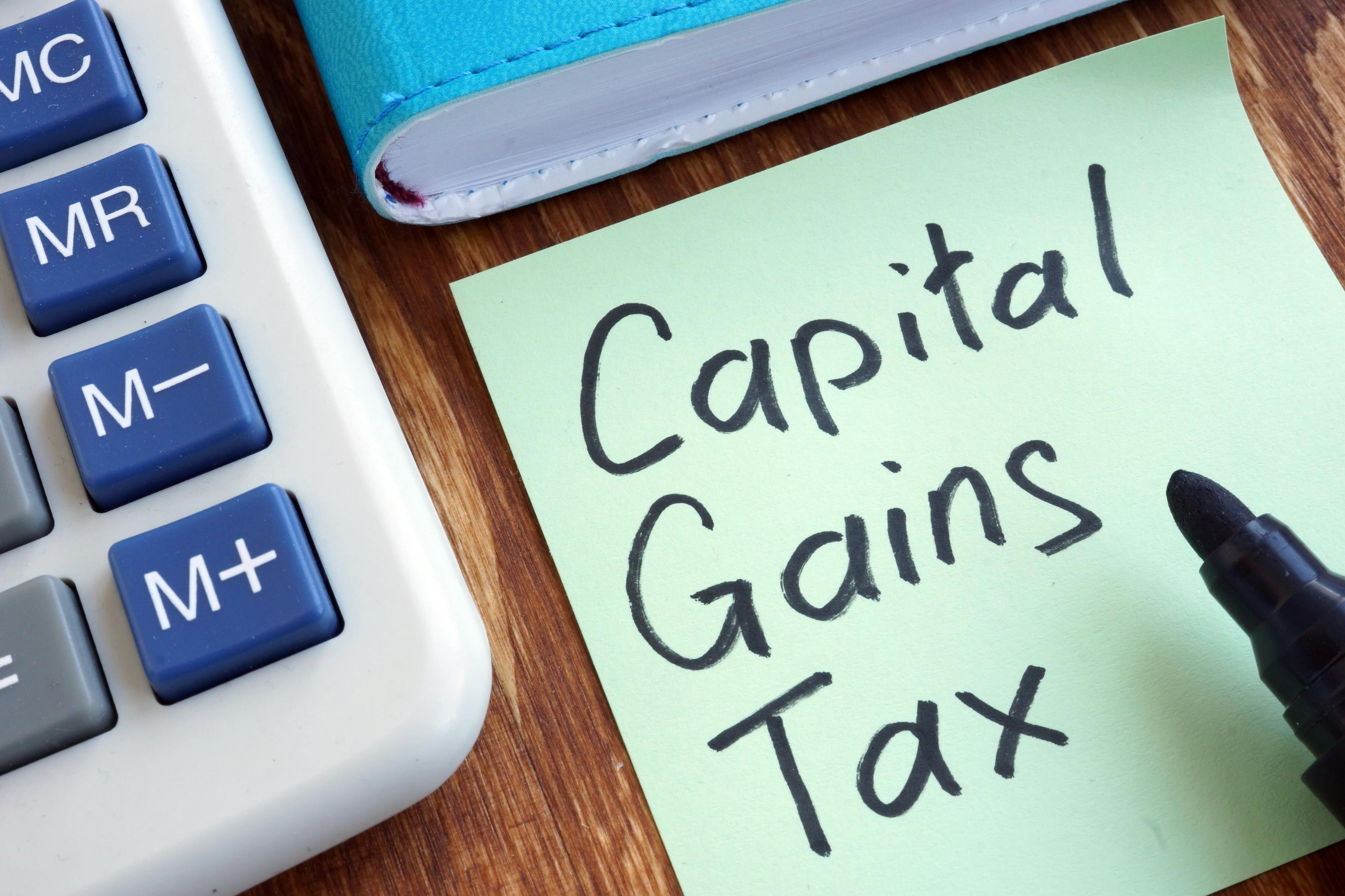 The image shows a portion of a calculator on the left with a post it note to the right with the words Capital Gains Tax written on it. There is the tip of a marker pen showing.  The image conveys How to Avoid Capital Gains Tax on Real Estate. 