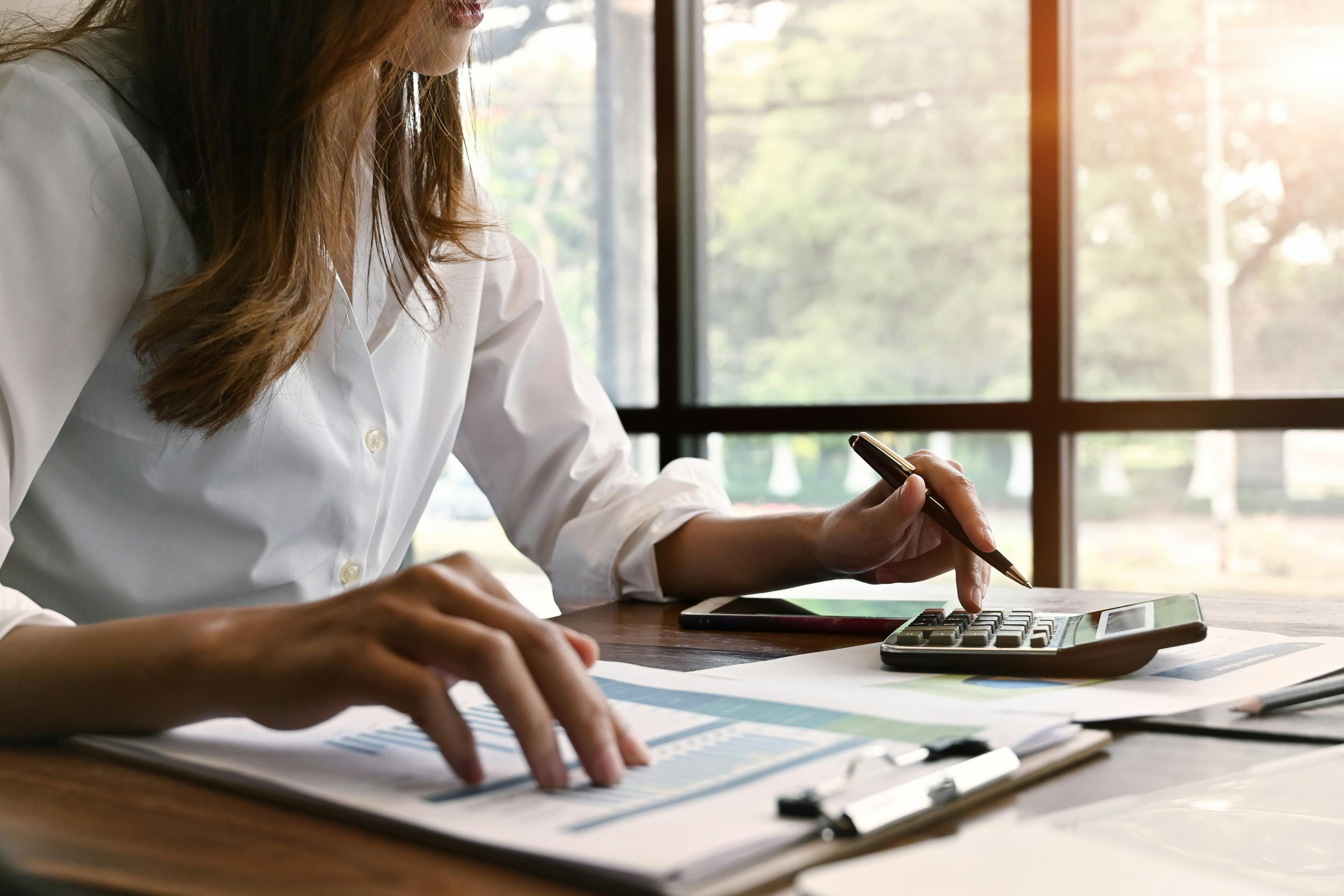 The image shows a woman wearing a white shirt sitting at a desk by a window. Her left hand is holding a pen, while she is using a calculator. Her left hand is on a clipboard with financial papers. This image conveys 5 Essential Property Management Bookkeeping Tips