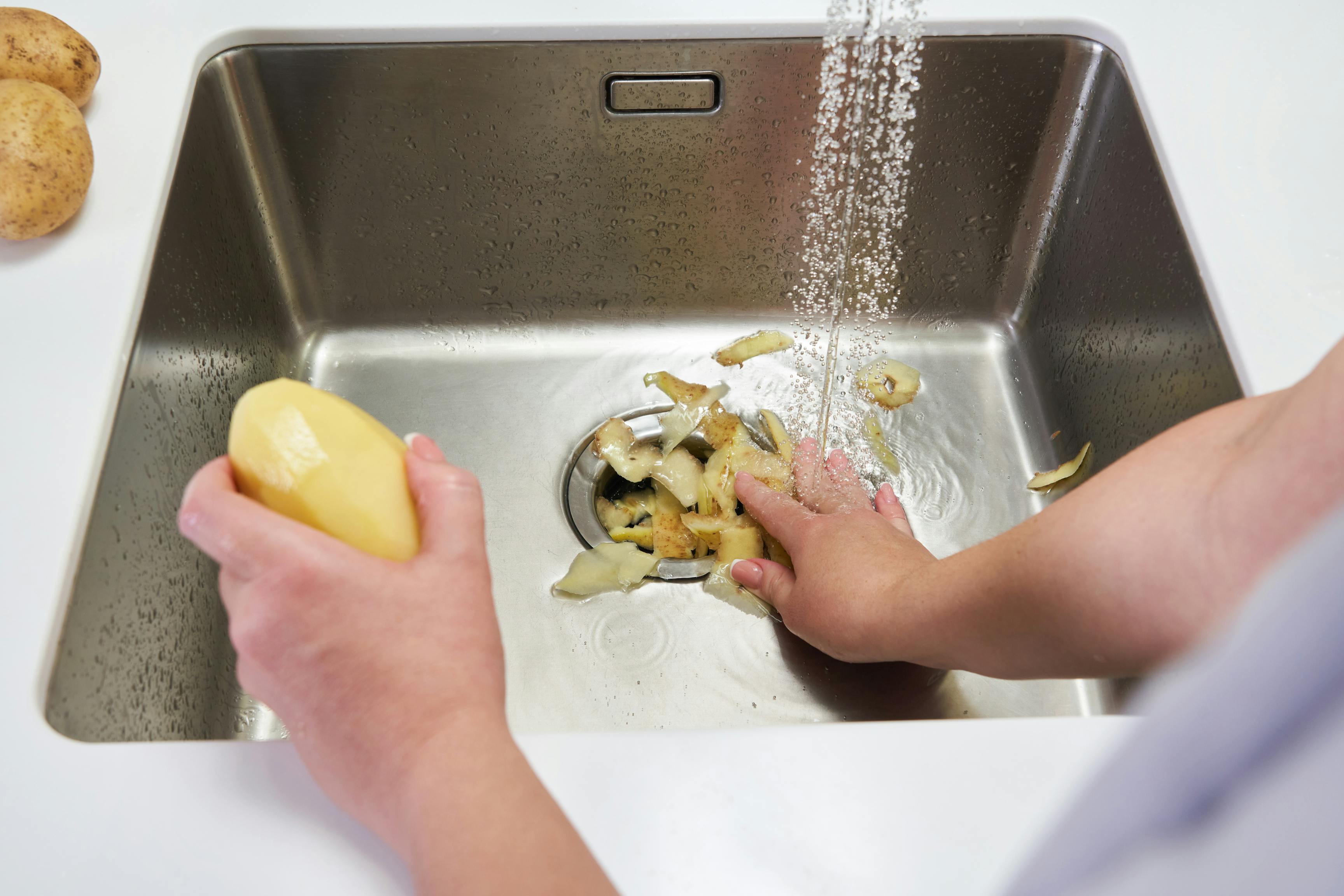 The image shows a white countertop with an inlaid stainless-steel kitchen sink. A woman’s right hand is holding a peeled potato, and her right hand is under a stream of water pushing the potato peels down the sink. To the left of the sink is a partial view of two unpeeled potatoes. This image conveys 9 things that can ruin your garbage disposal, according to plumbers.
