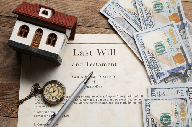 The image shows a Will on a desk. On one side is a small model home next to a pocket watch and a pen. On the other side are $100 dollar bills. This image conveys Estate Planning for Rental Housing Owners