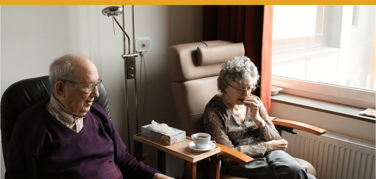 Older man and older woman seated in comfortable chairs by a window, with a cup of coffee and tissues on a small table between them.