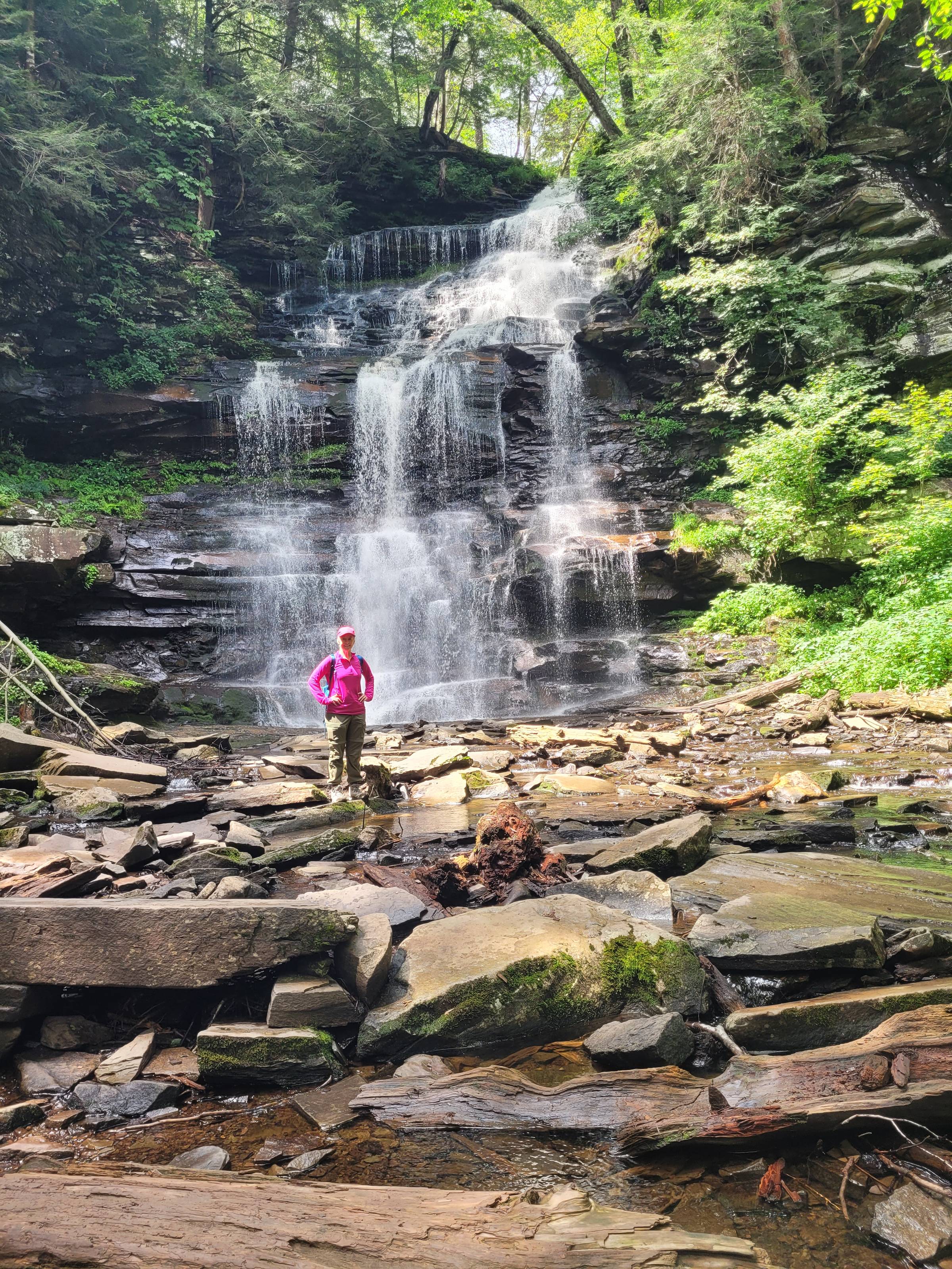 A person in a bright pink jacket and olive-green pants stands confidently on a rocky, shallow stream bed at the base of a tall, tiered waterfall. The waterfall cascades down dark, moss-covered rock ledges surrounded by dense, green forest. Sunlight filter