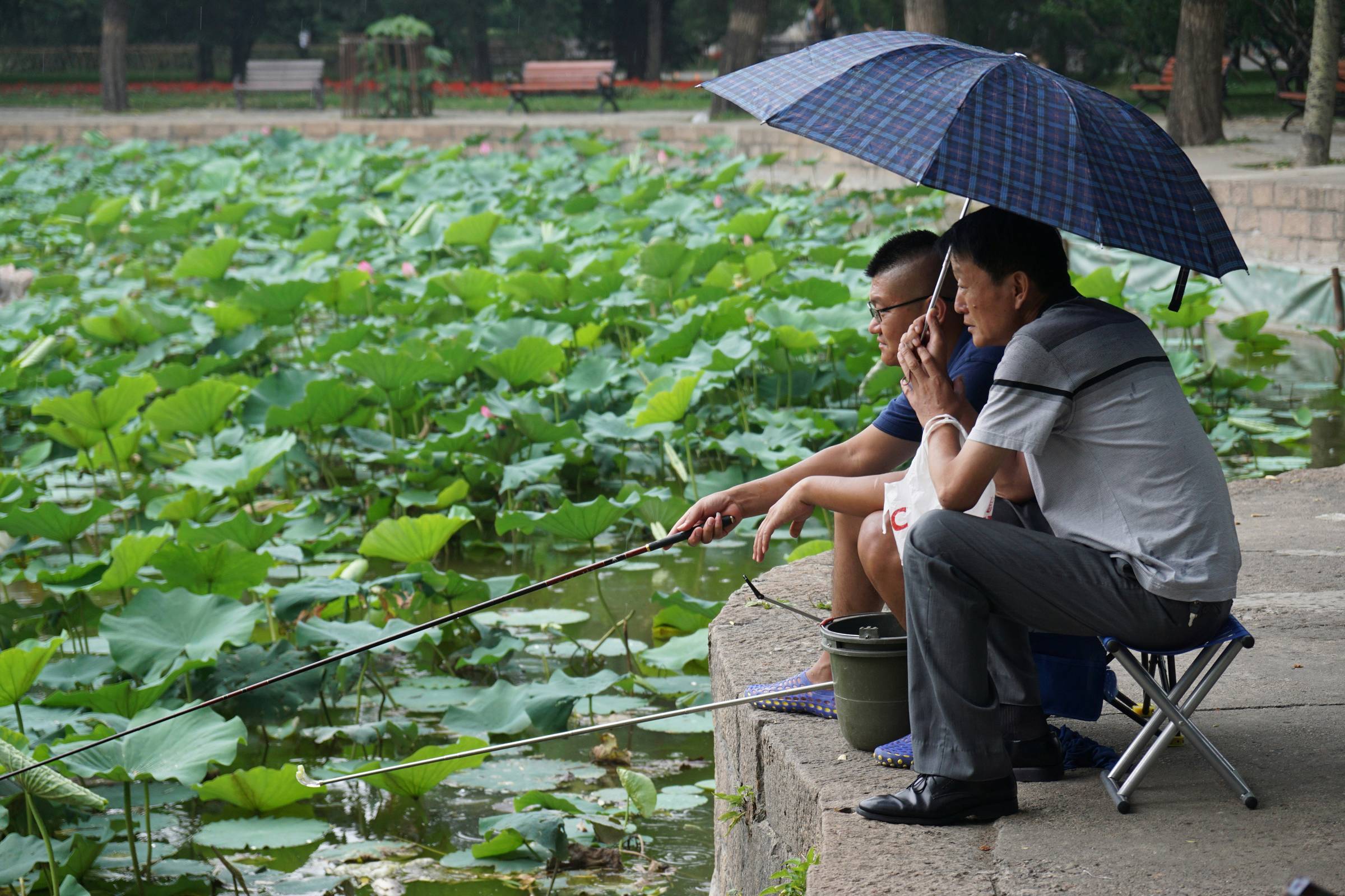 One man holds an umbrella over another man who is fishing