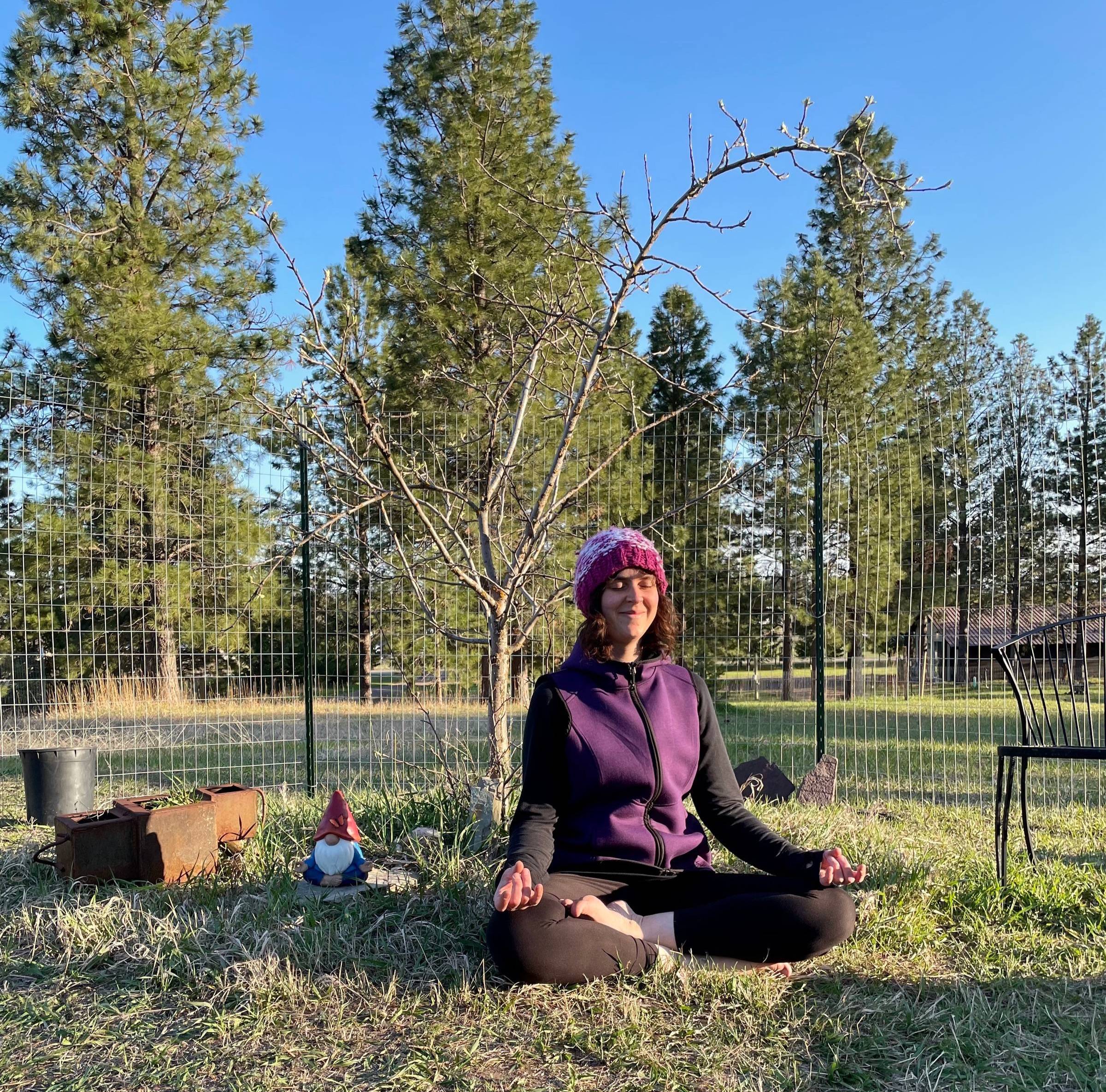 Photo of life coach Kelsey Holmes sitting in meditation under a budding apple tree, next to a meditating garden nome.