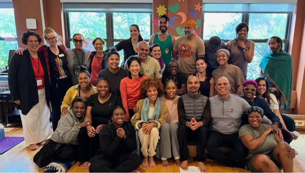 A group of yoga teacher training graduates stands together, arranged in a loose cluster and facing the camera. The setting appears natural, with greenery and open space in the background. The group includes multiple people standing close together, some wi
