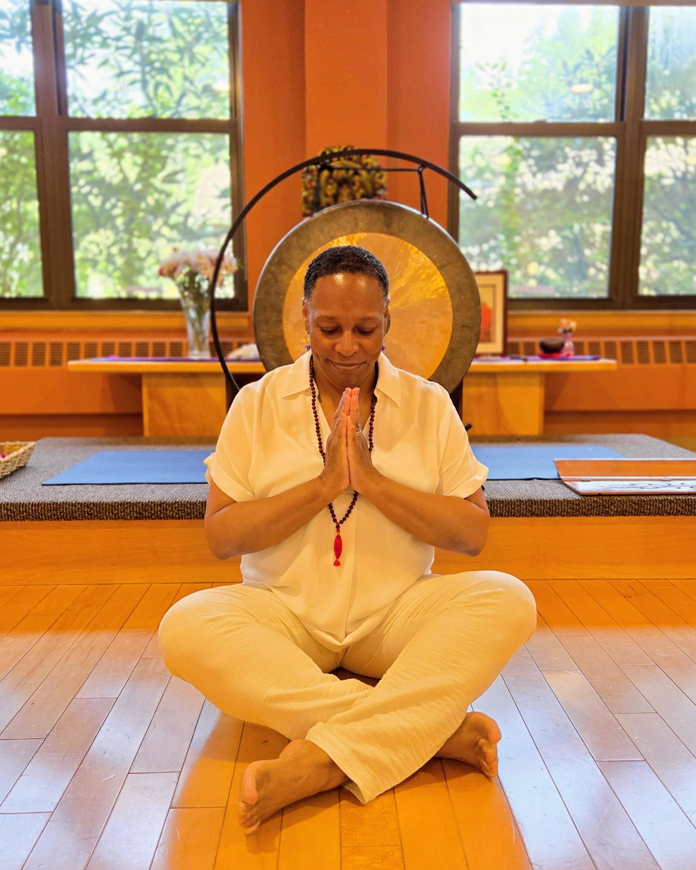 Person sits on the ground on a mat, wearing light-colored clothing, with a calm and grounded posture. The setting is simple and uncluttered, with soft natural lighting. The body is upright but relaxed, suggesting stillness and ease. The overall composition feels quiet and spacious, visually supporting themes of rest and gentle awareness.