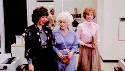 Lily Tomlin wearing blue and black, Dolly Parton wearing a powder blue dress, and Jane Fonda wearing a pink blouse and skirt walk through their office in the movie 9-to-5, whispering and laughing with one another. 