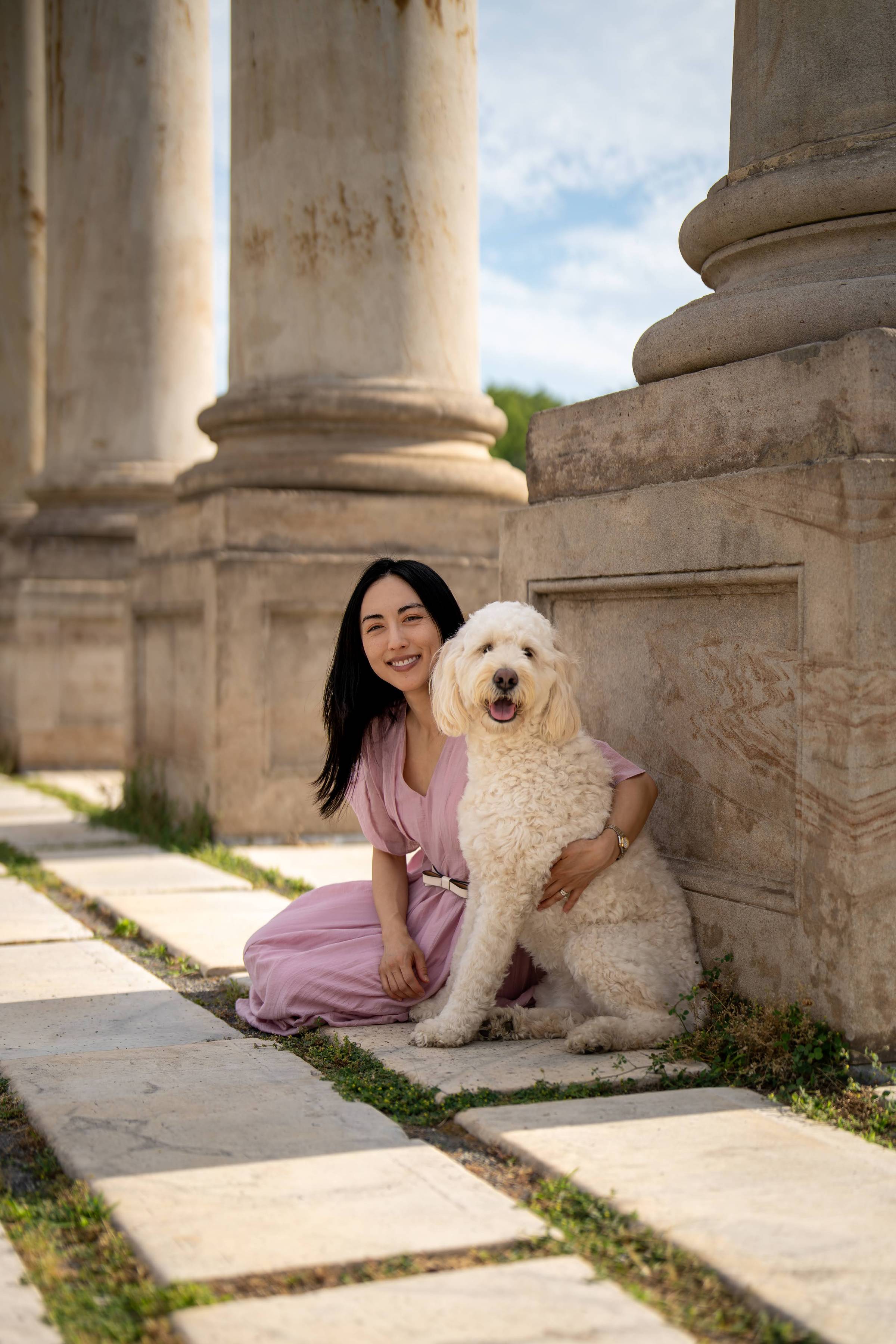 A smiling woman with long dark hair, wearing a pink dress with a white belt, kneels beside a large cream-colored Goldendoodle on marble paving stones between weathered stone columns at an ancient ruin site.