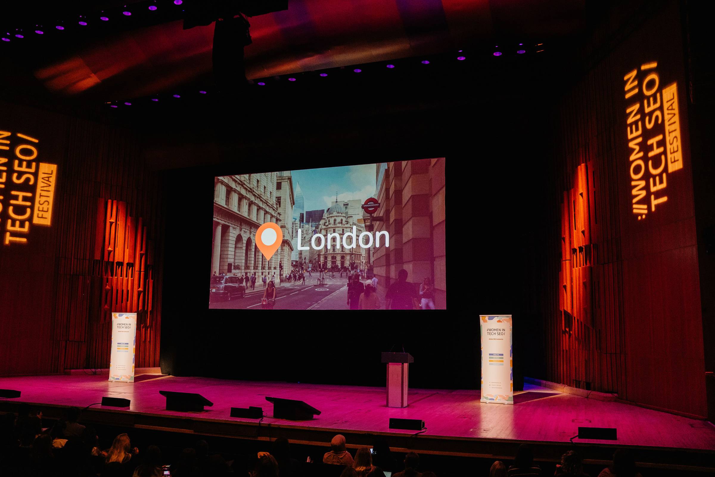 Wide photo of the WTSFest conference stage before a session begins. A large screen displays an image of London with a location pin, while Women in Tech SEO Festival branding is projected on tall banners at either side of the stage. A podium stands in the centre, with audience members seated in the foreground.
