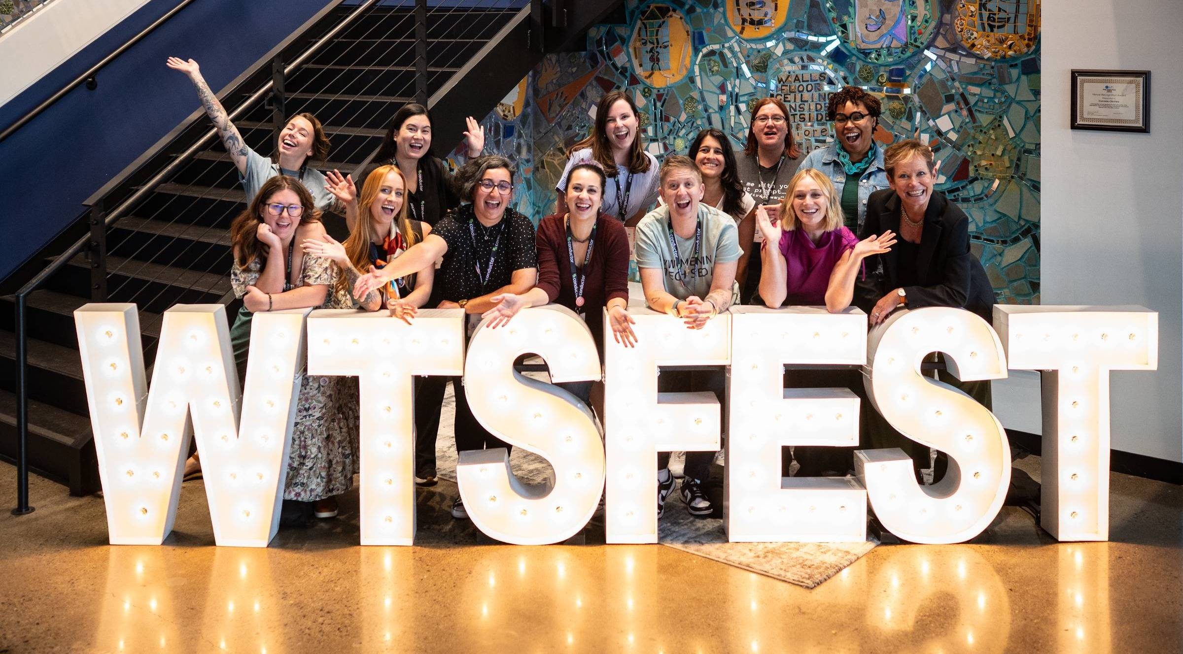 Group photo of eleven Women in Tech SEO attendees gathered behind large illuminated “WTSFEST” letters inside a venue. Several people wave or raise their arms, smiling in front of a colourful mosaic wall and staircase.