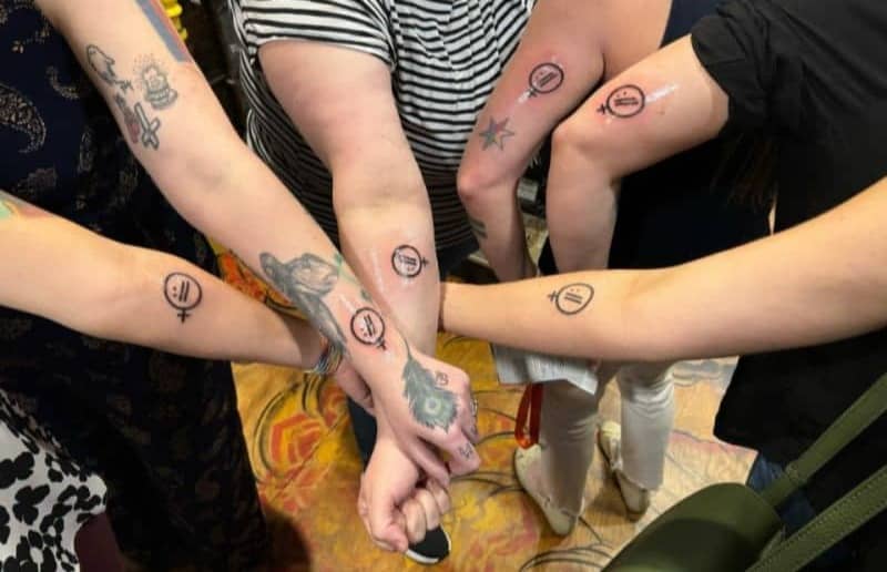 Six people stand in a circle with their forearms extended towards the centre, showing matching Women in Tech SEO logo tattoos on their arms. Several participants have additional tattoos, and all of the logo tattoos are visible together as a shared keepsake from the event.