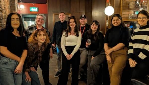 Group photo of attendees from a Merseysearch event standing together in a bar-style venue with warm lighting. Around ten people pose and smile at the camera, dressed casually, with wooden interiors and hanging lights visible in the background.