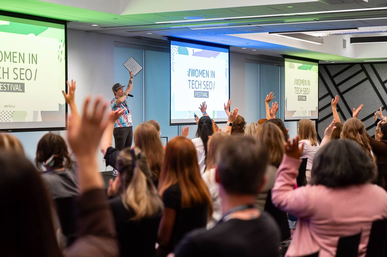 Audience members raise their hands during an interactive session at WTSFest Philadelphia. A speaker wearing a colourful shirt and cap stands at the front holding a microphone and raising a clipboard. Three large screens display the Women in Tech SEO Festival logo.