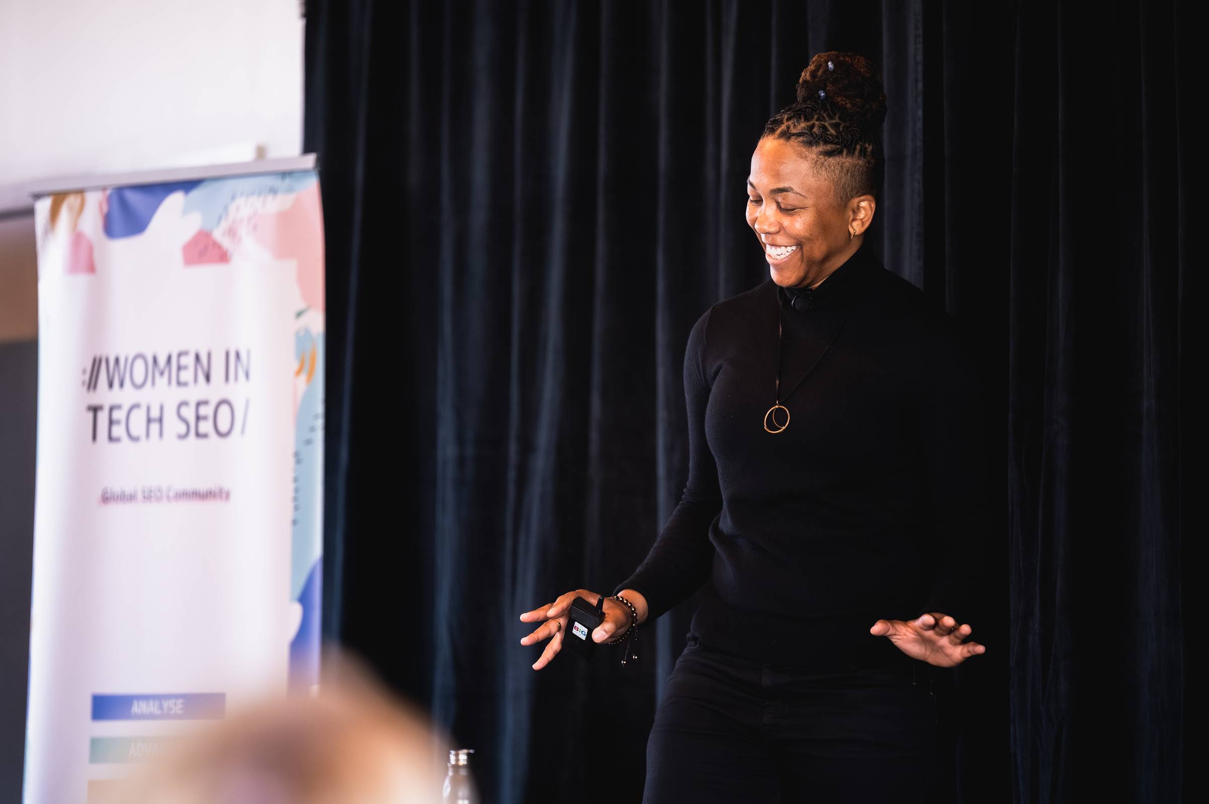 A smiling speaker dressed in black presents on stage beside a Women in Tech SEO banner, gesturing to the audience against a dark curtain backdrop.
