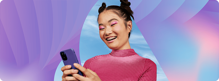 Smiling young woman with space buns hairstyle and pink eye makeup looking at a smartphone in her hands. She is wearing a pink patterned top and braces, standing against a colorful gradient background with soft purple and blue tones.