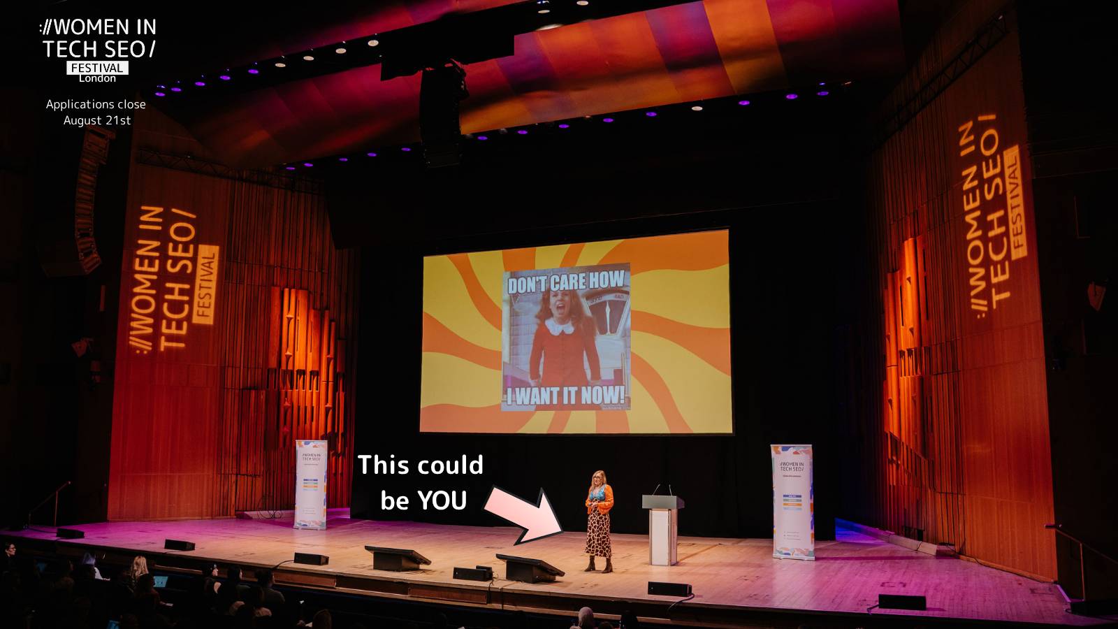 Wide view of the WTSFest stage inside a large auditorium, with a speaker presenting beneath a giant projection screen. An arrow points to the speaker with the words “This could be YOU,” encouraging future speaker applications. Women in Tech SEO branding is displayed on both sides of the stage.