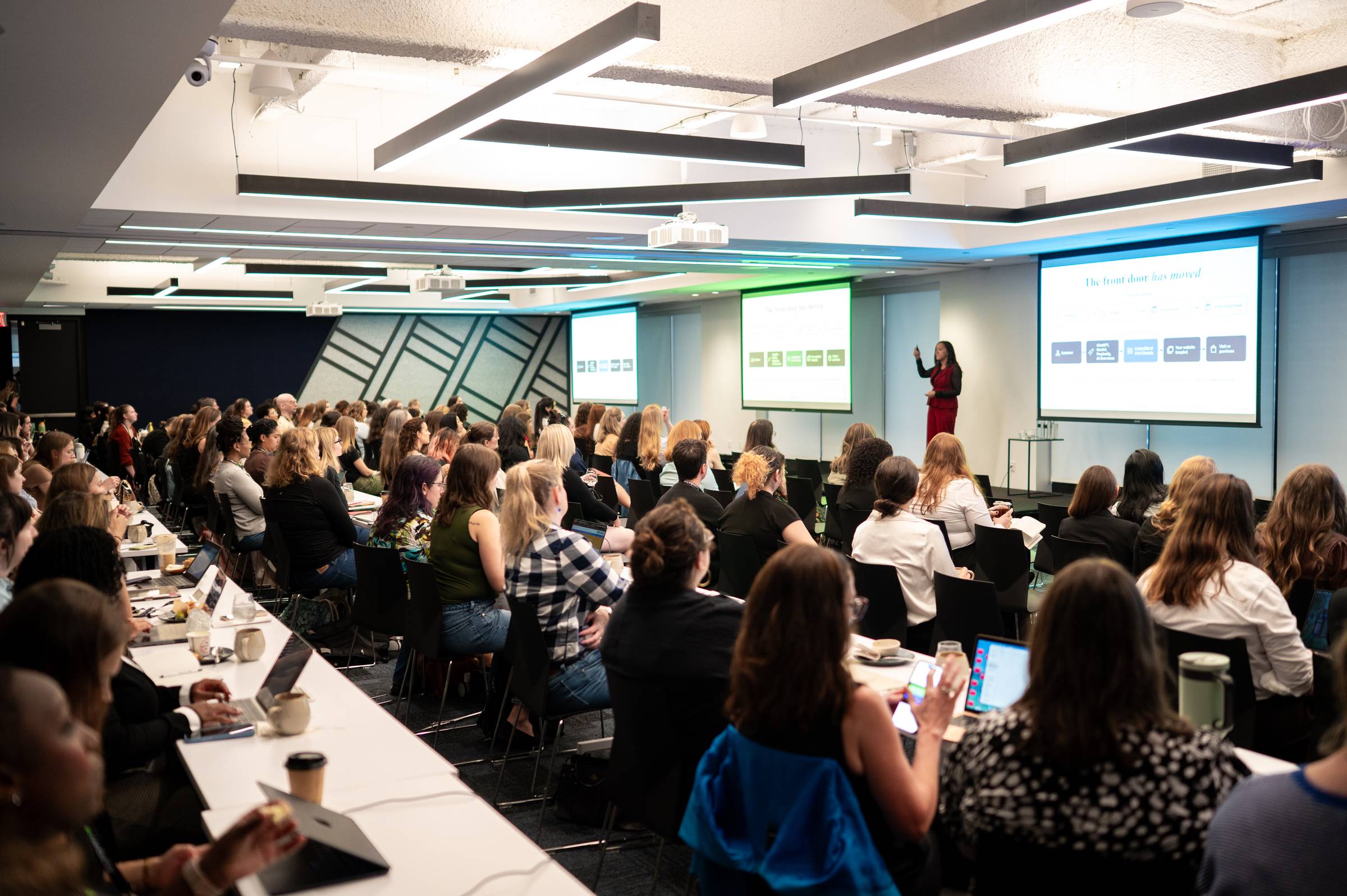 Wide-angle photograph of Krystal Taing speaking on stage at WTSFest Philadelphia. She wears a red outfit with a black jacket and gestures towards a presentation displayed on three large screens. The slide reads “The front door has moved.” A large audience, predominantly women, sits in rows facing the stage, with some attendees taking notes or using laptops. The modern conference room has long white tables, geometric wall panels and angular overhead lighting.
