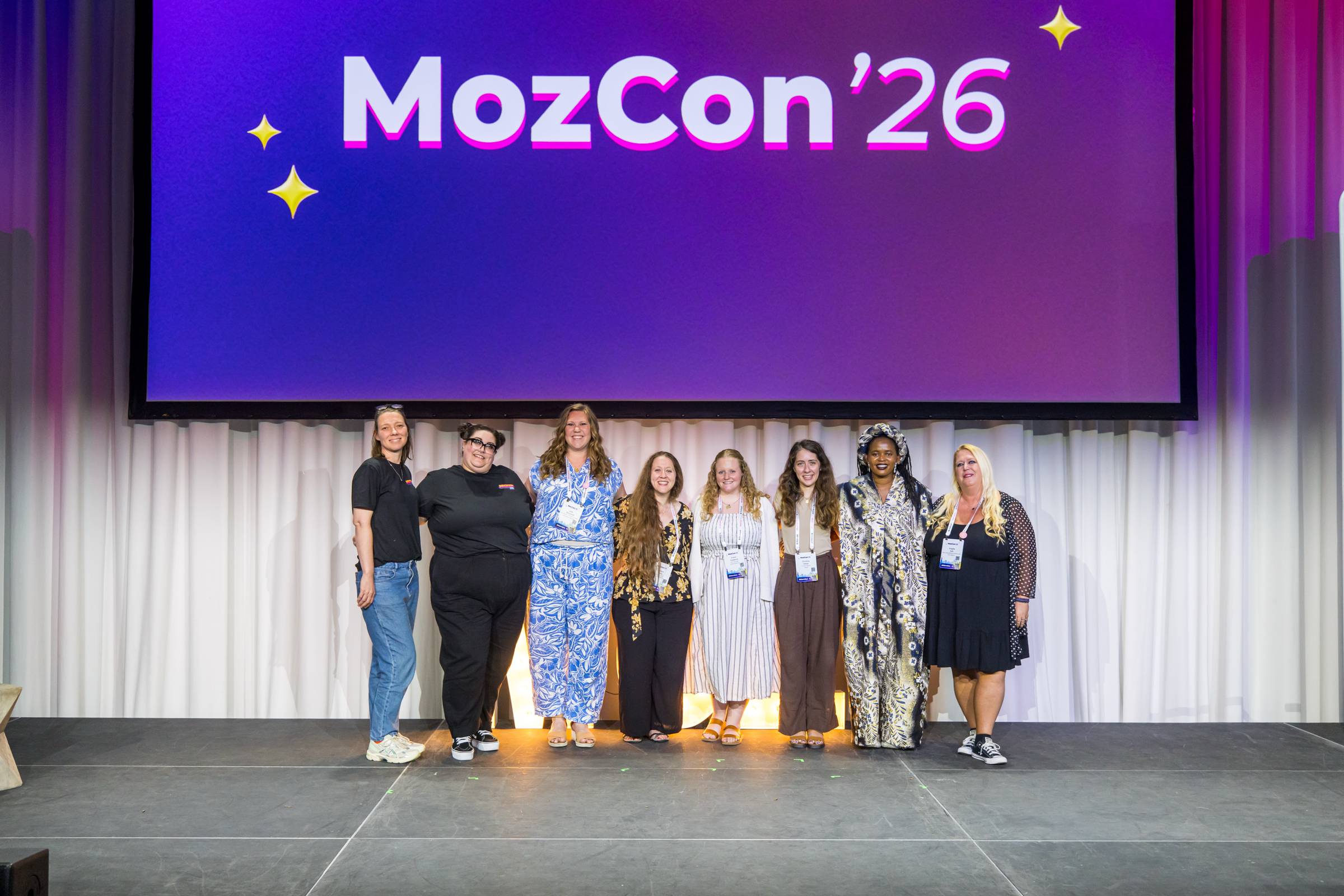 Group photo of eight conference attendees standing on stage beneath a large screen displaying “MozCon ’26.” Everyone is smiling towards the camera, wearing event badges after the conference.