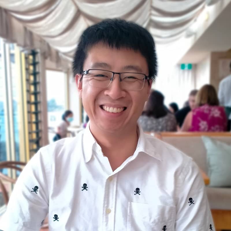 A picture of Paul smiling. Paul is an Australian born Vietnamese web developer. He has black hair, wears glasses and is wearing a white short sleeved button up top with skulls on them.