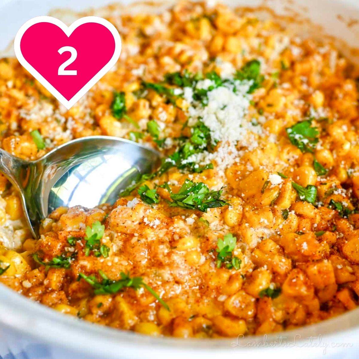 A close-up of a creamy, cheesy Mexican street corn dip or casserole in a white dish, with a large metal serving spoon scooping a portion out. The corn is coated in a reddish spice blend and garnished with crumbled cotija cheese and fresh chopped cilantro. A pink heart icon with the number 2 is in the upper left.