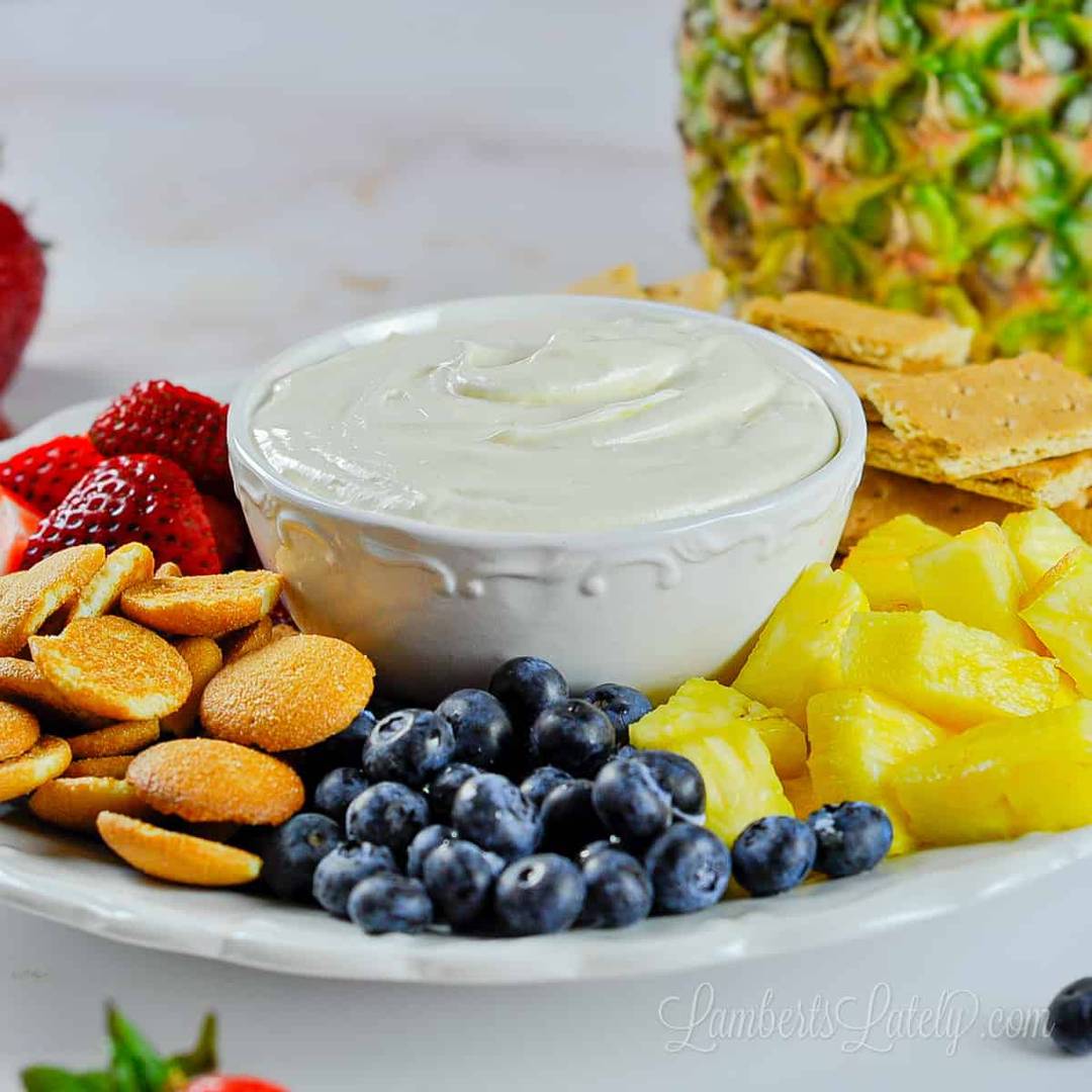A white serving platter arranged with a small bowl of smooth fruit dip surrounded by fresh strawberries, blueberries, pineapple chunks, graham crackers, and vanilla wafer cookies.