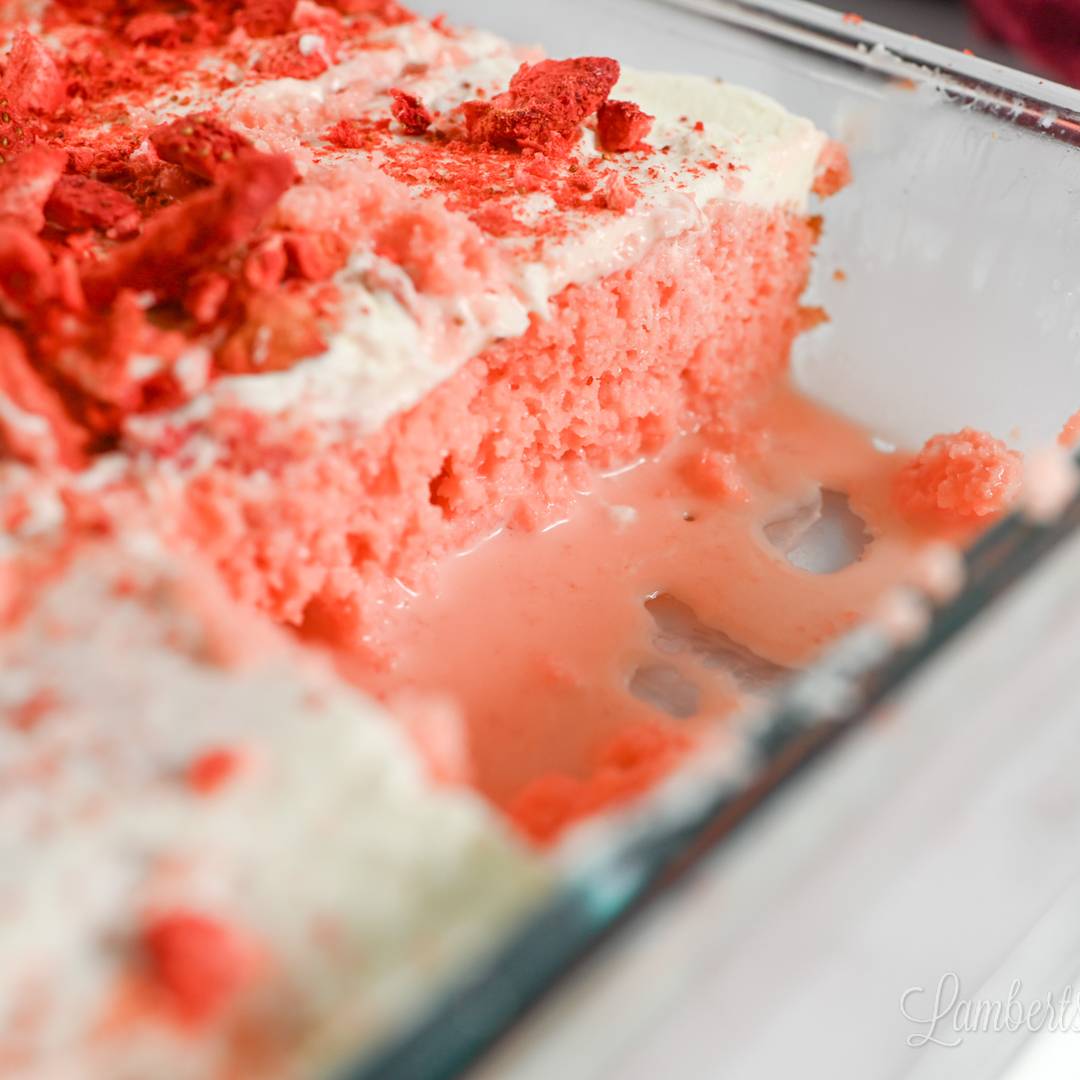A close-up side view of a strawberry poke cake in a glass baking dish, featuring a bright pink sponge soaked in glaze and topped with creamy white frosting and crushed freeze-dried strawberries.