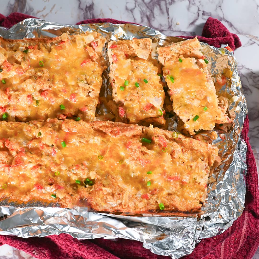 Crawfish bread topped with creamy crawfish filling, melted cheese, and chopped chives on a foil-lined baking sheet.