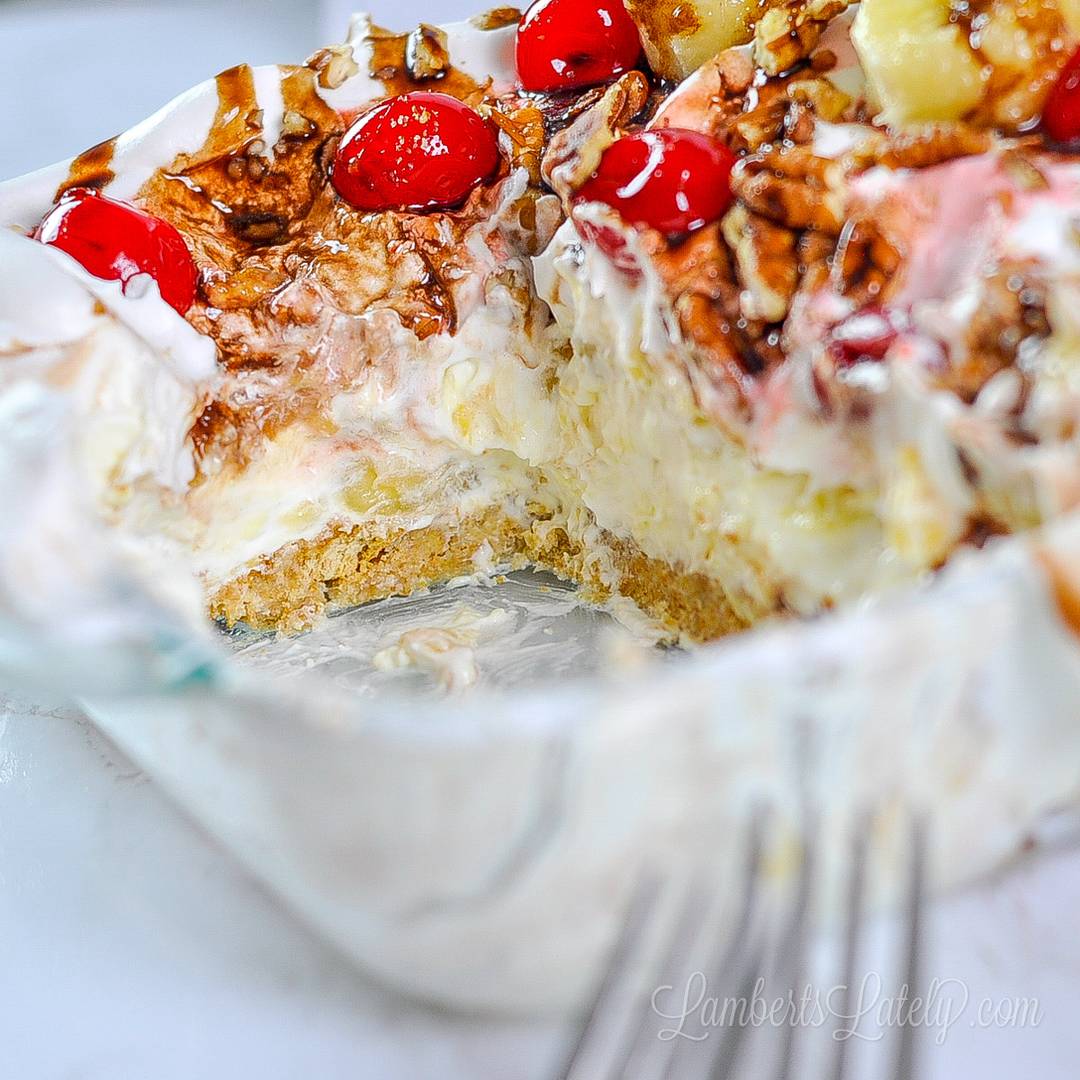 A cross-section of a layered banana split dessert in a glass dish, featuring a graham cracker crust, a creamy pineapple layer, whipped topping, chocolate drizzle, chopped pecans, and maraschino cherries.