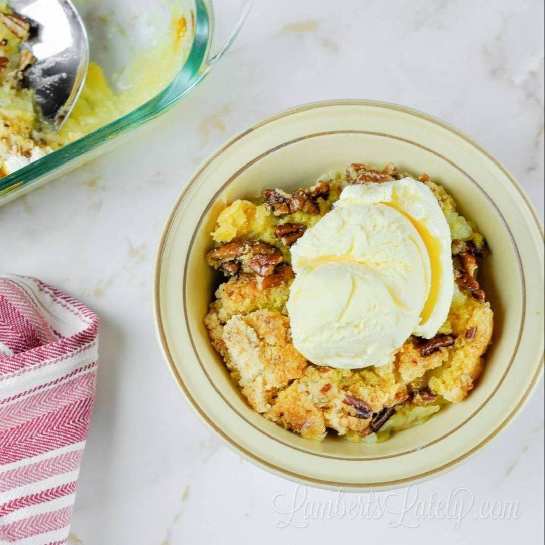 A top-down view of a warm serving of pineapple dump cake in a yellow bowl, topped with a large scoop of melting vanilla ice cream and crunchy pecans.