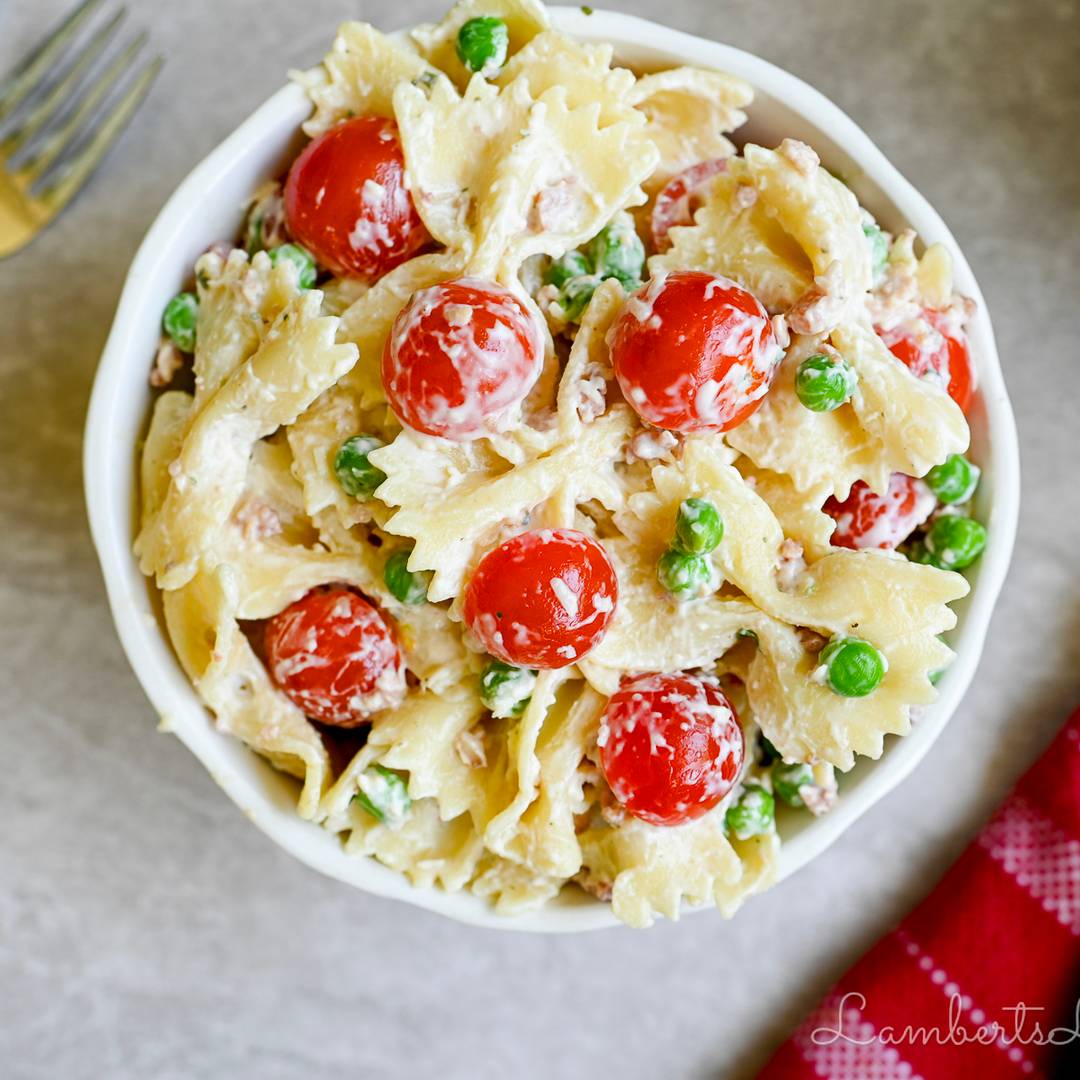 A top-down view of a creamy bow-tie pasta salad in a white bowl, tossed with bright red cherry tomatoes and green peas, with a red patterned napkin in the corner.