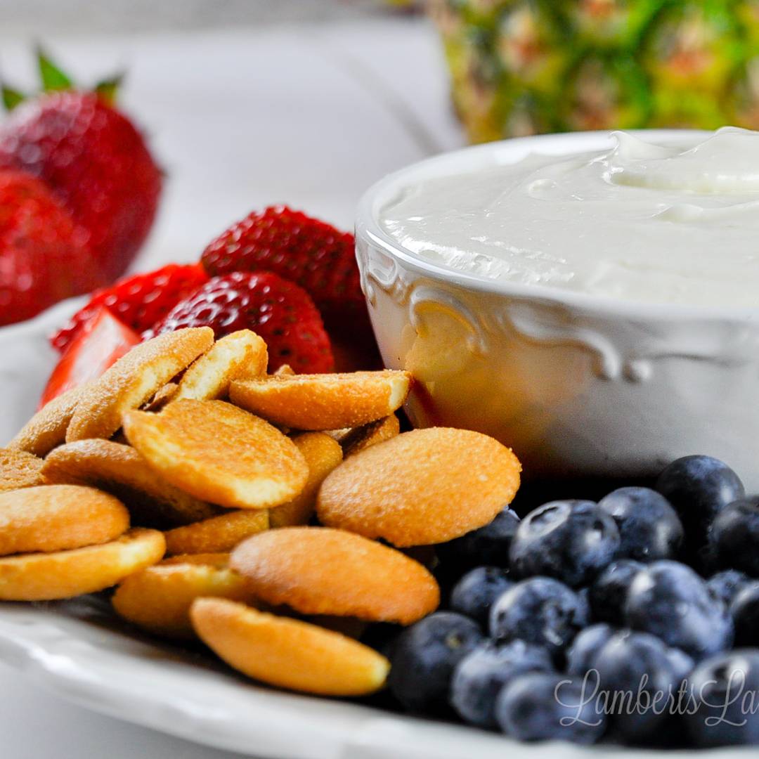 A white bowl of fluffy cream cheese fruit dip served on a platter with fresh strawberries, blueberries, and vanilla wafer cookies.