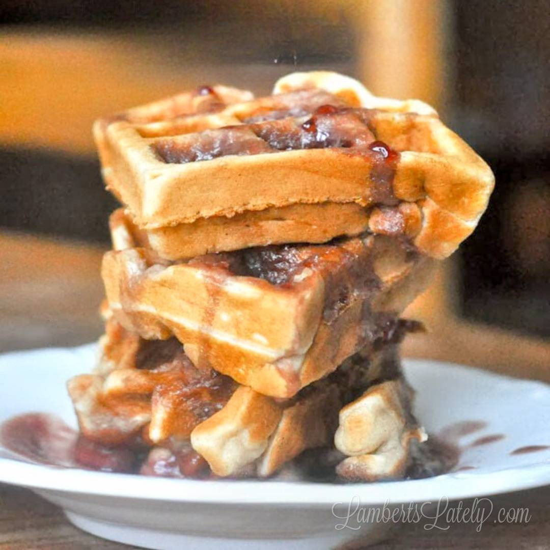 Golden waffles stacked and topped with a warm syrup or fruit compote, shown on a white plate with sauce dripping down the sides.