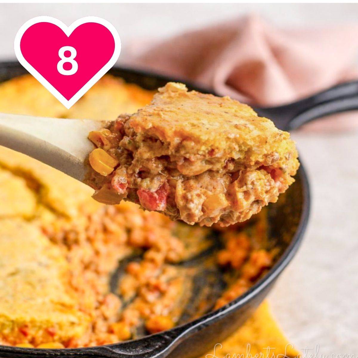 A close-up shot of a wooden spoonful lifting a large, thick portion of a cheesy beef and corn casserole topped with a golden cornbread crust. The rest of the dish is visible inside a black cast-iron skillet in the background. A pink heart icon with the number 8 is in the upper left.