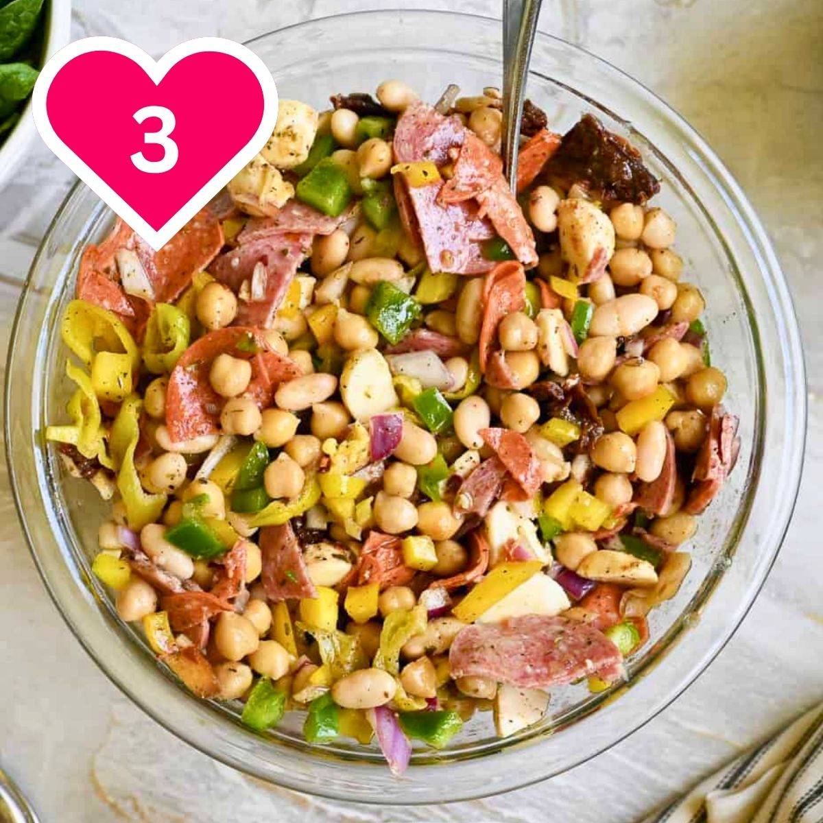 An overhead shot of a glass mixing bowl filled with an Italian-style chopped salad. The salad features chickpeas, white beans, slices of pepperoni and salami, green and yellow bell peppers, red onion, and fresh mozzarella tossed in an herb dressing. A pink heart icon with the number 3 is in the upper left.