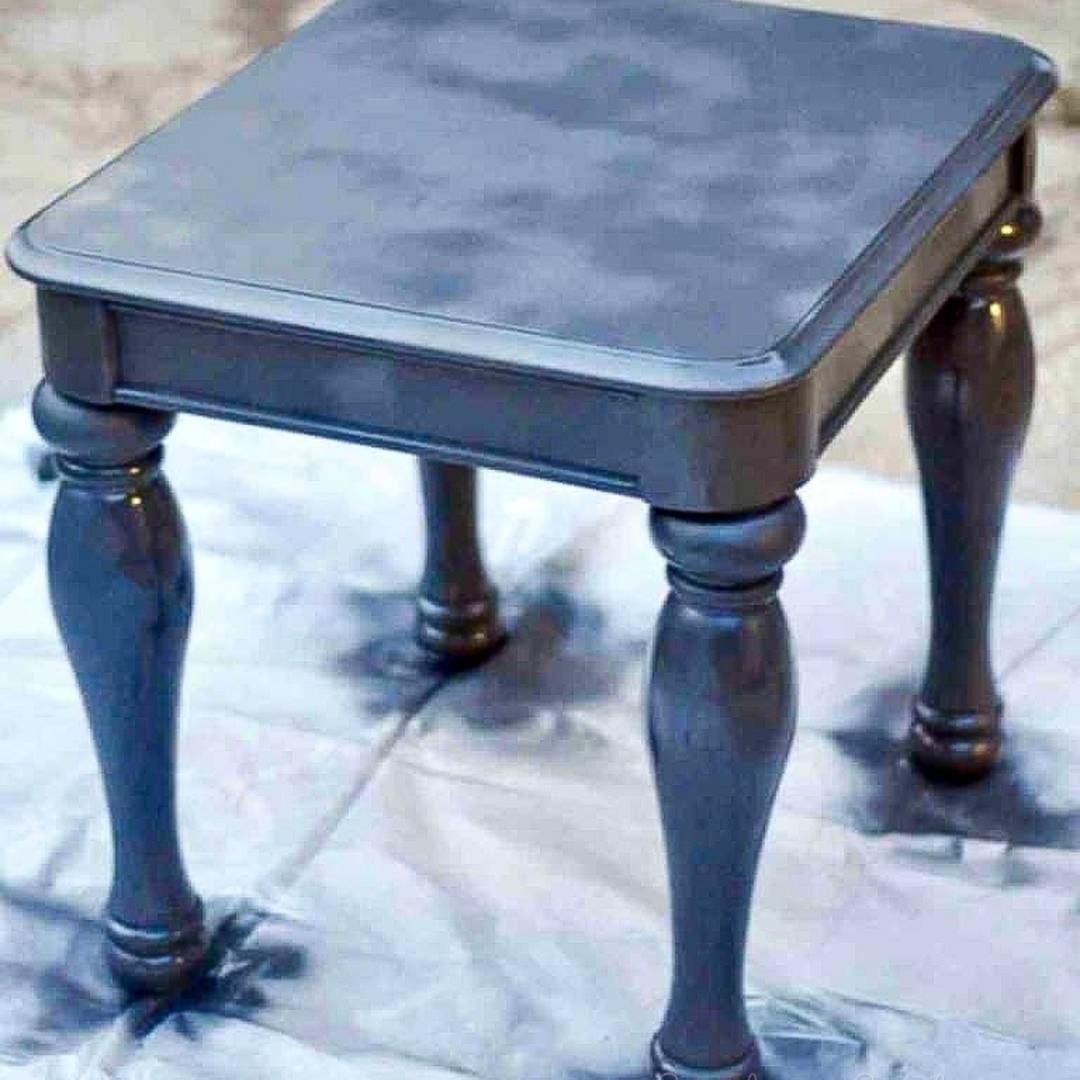 A small, square wooden side table with decorative turned legs being spray-painted a dark navy blue on top of a drop cloth.