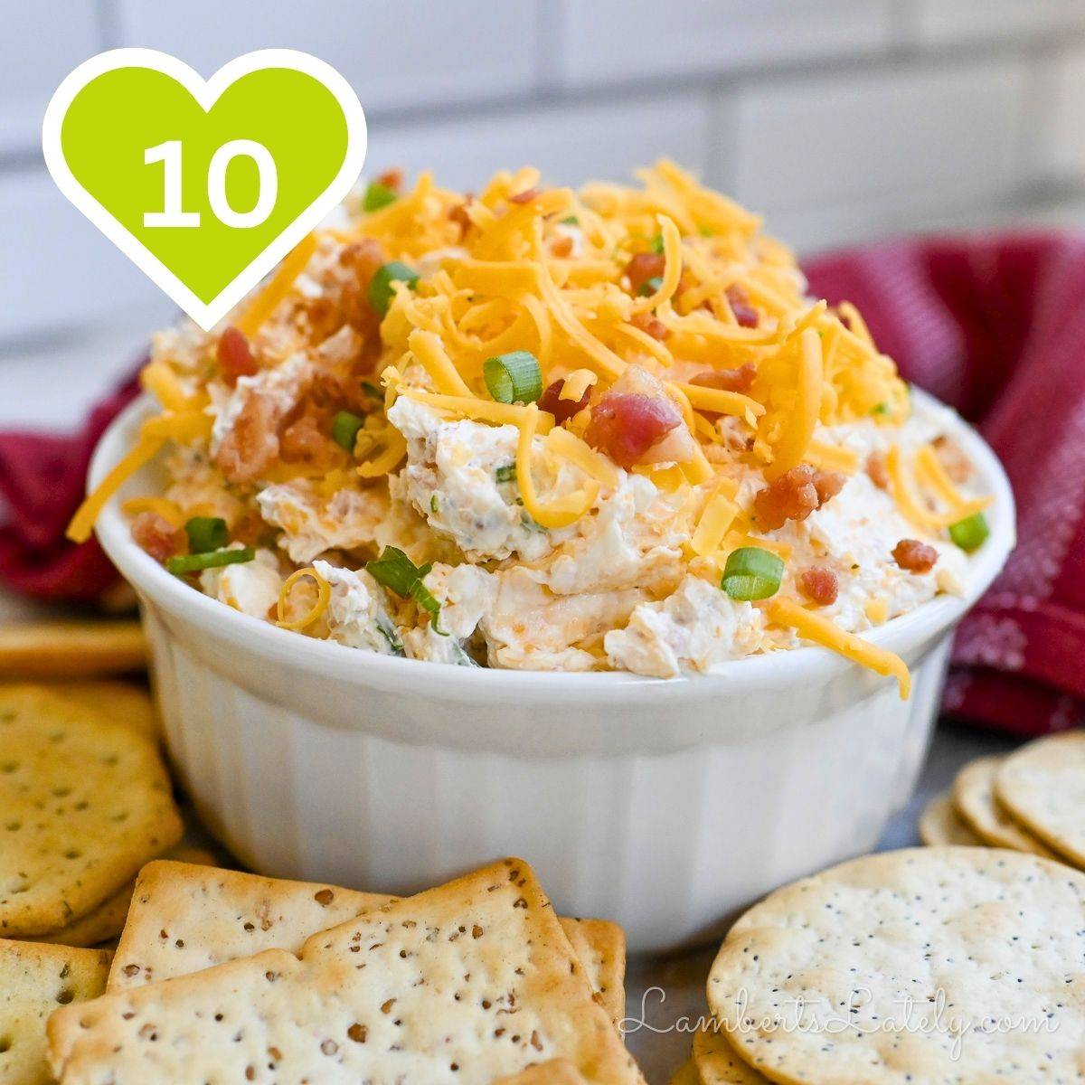 Creamy party dip topped with shredded cheese and green onions in a white bowl, served with crackers.