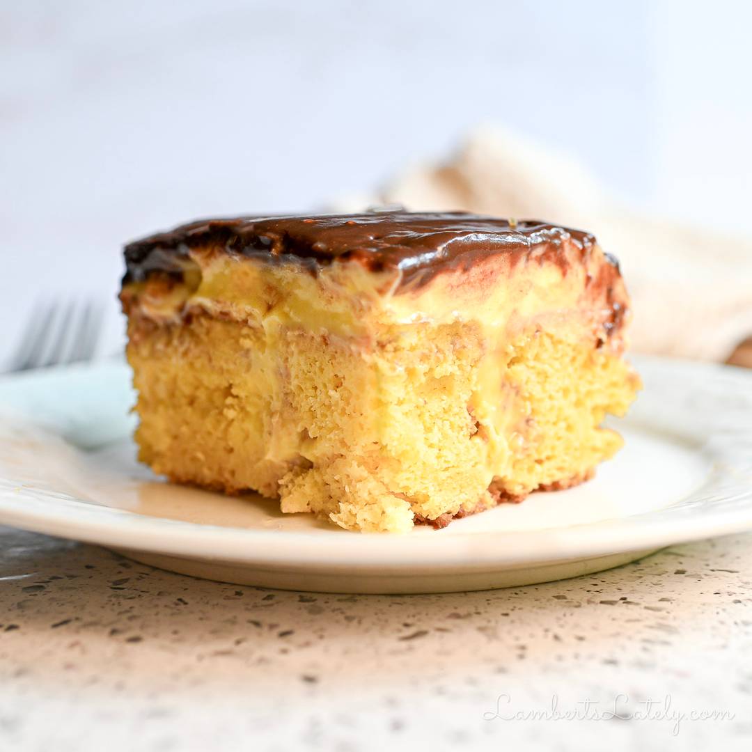 A large slice of Boston cream poke cake on a white plate, showing the yellow cake filled with vanilla pudding and topped with a thick layer of chocolate ganache.