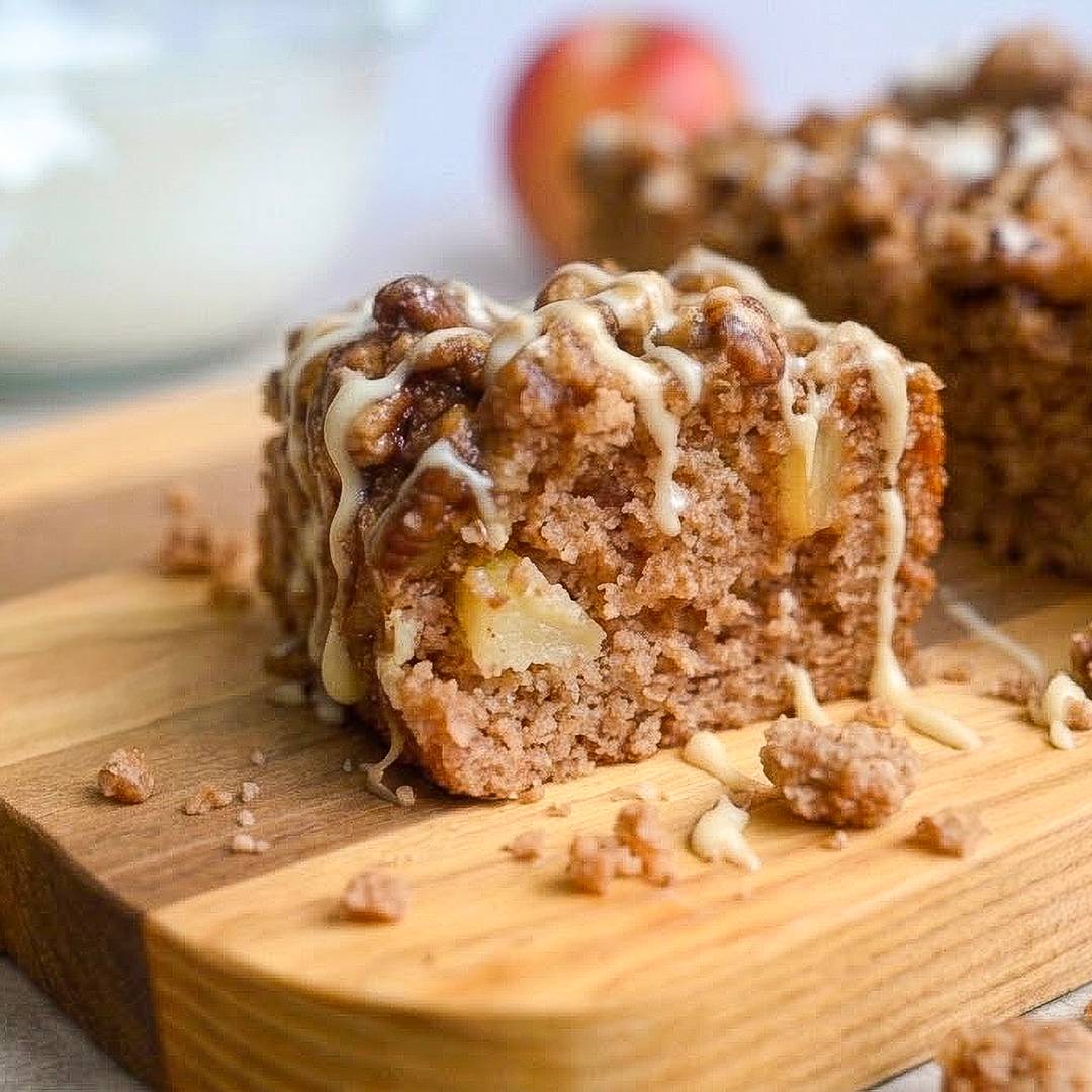 Slice of apple spice cake with crumb topping and glaze on a wooden board.