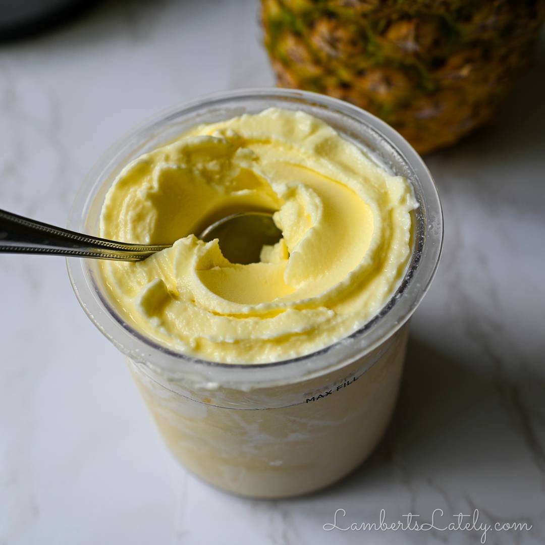A Ninja Creami pint of pale yellow Dole Whip with a deep swirl from a spoon, positioned next to a fresh pineapple.