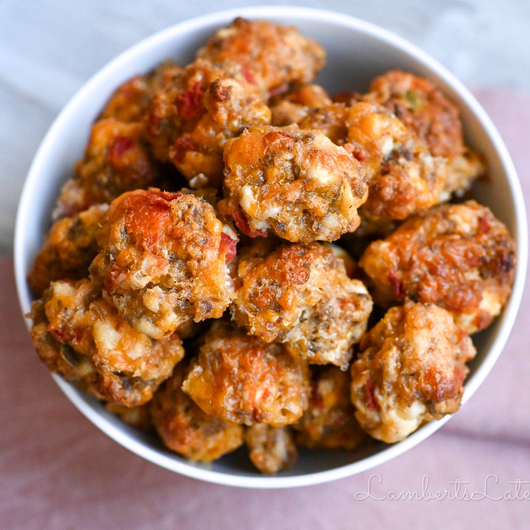 A close-up, overhead shot of a white bowl filled with golden-brown, cheesy sausage balls, showing a visible texture of melted cheese and herbs.